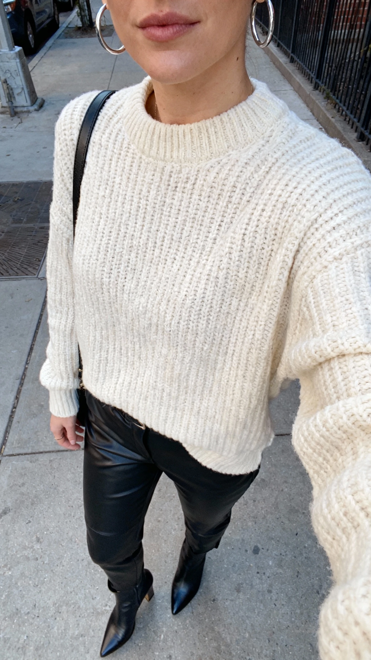 iPhone street-side selfie cropped at shoulders showing a woman in a chunky cream sweater with black leather (or faux) pants, heeled ankle boots, silver hoops, and a crossbody, face not visible, city sidewalk, natural daylight, casual iPhone photo.