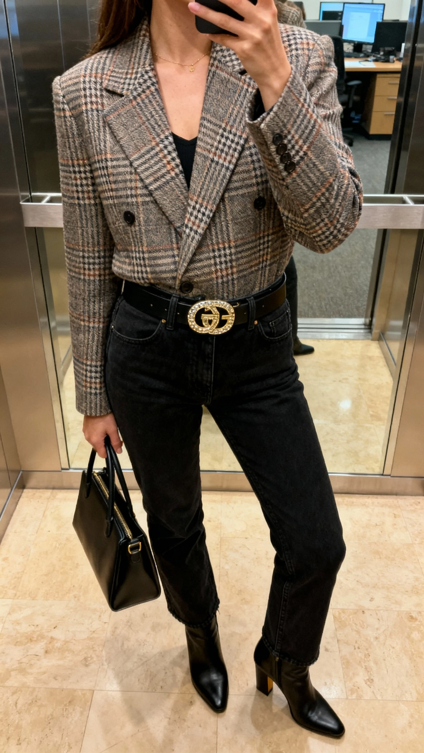 iPhone waist-up selfie cropped at neck of a woman wearing a plaid blazer, black jeans, statement belt with bold buckle, black heeled boots, and structured tote; face not visible, office elevator reflection, warm indoor lighting, natural iPhone photo quality.