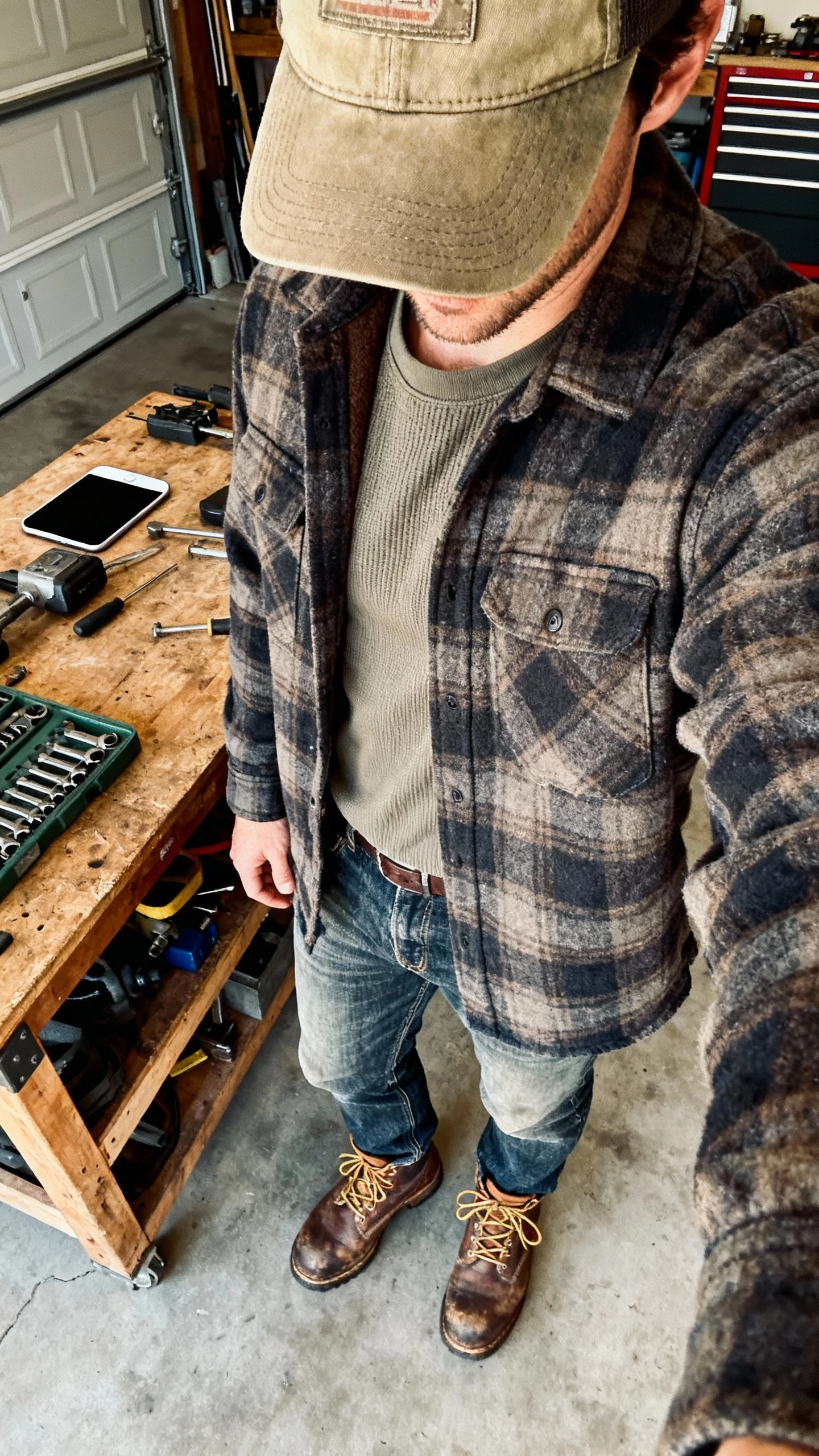 iPhone workbench selfie from neck down of a man wearing a heavyweight plaid flannel over a thermal tee, rugged straight-leg denim, a canvas work cap, and lace-up work boots, face not visible, garage setting with natural side light, natural iPhone quality.