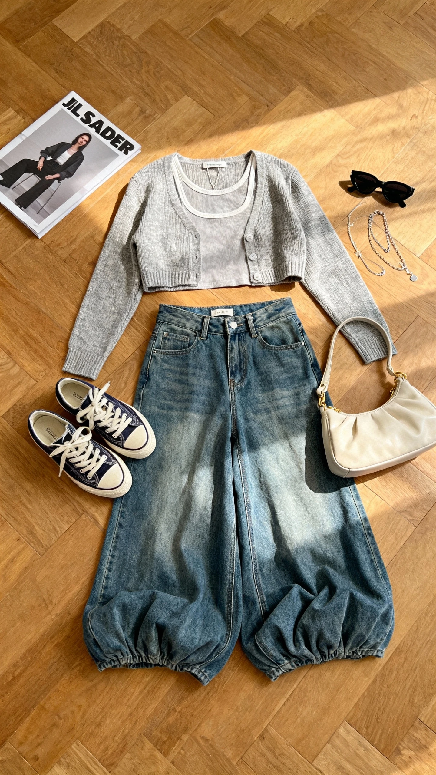 Top-down flat lay of cropped cardigan layered over a simple tank, puddle-hem wide-leg denim, retro sneakers, baguette bag, black sunglasses, and delicate layered necklaces on a wooden floor with a fashion magazine as prop, bright natural light, no person visible, clean organized composition, iPhone photo aesthetic.