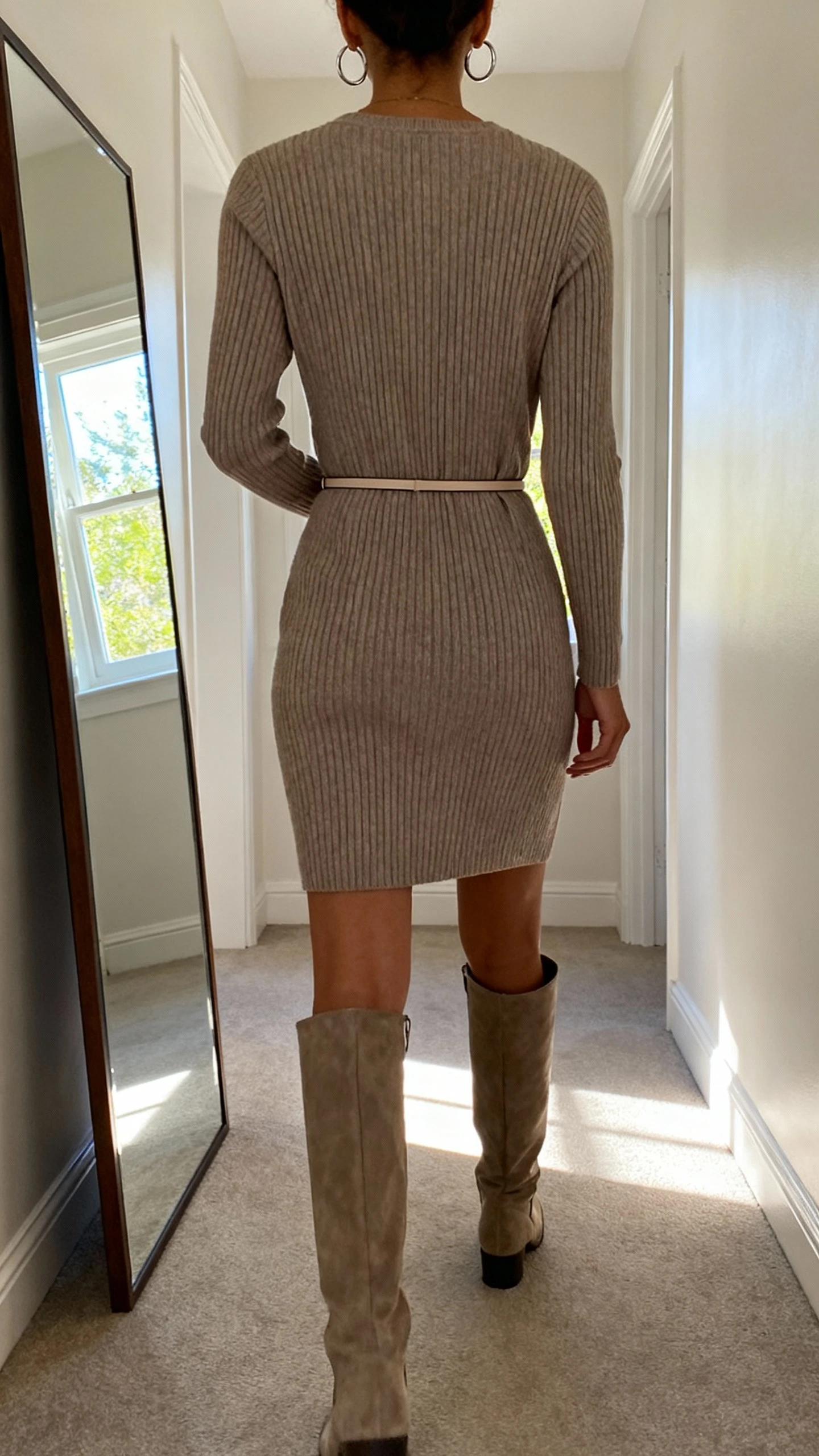 iPhone back view of a woman wearing a ribbed knit dress with tall knee-high boots, thin waist belt, and simple hoop earrings, face not visible, hallway full-length mirror, natural window light, iPhone photo quality.