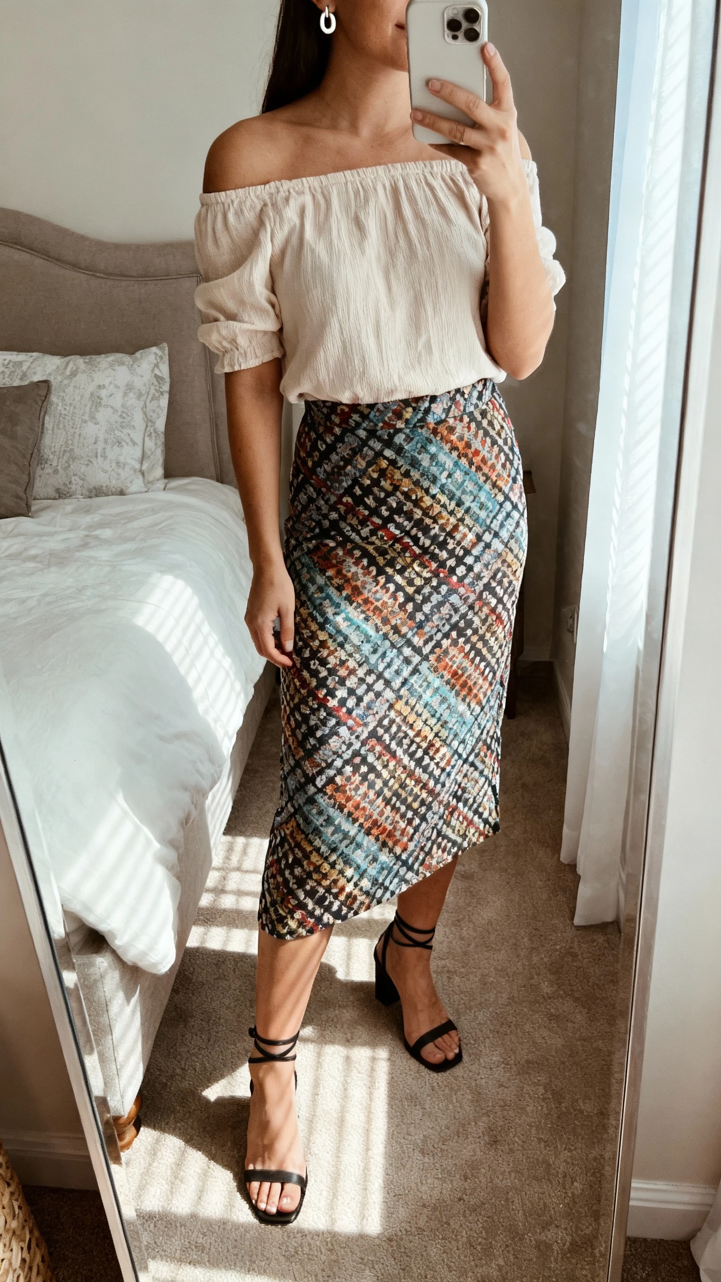 iPhone mirror selfie of a woman in a statement skirt paired with an off-shoulder top, ankle-strap heels, and minimalist earrings, face not visible, cropped at neck, bedroom mirror, diffused window light, iPhone photo quality.