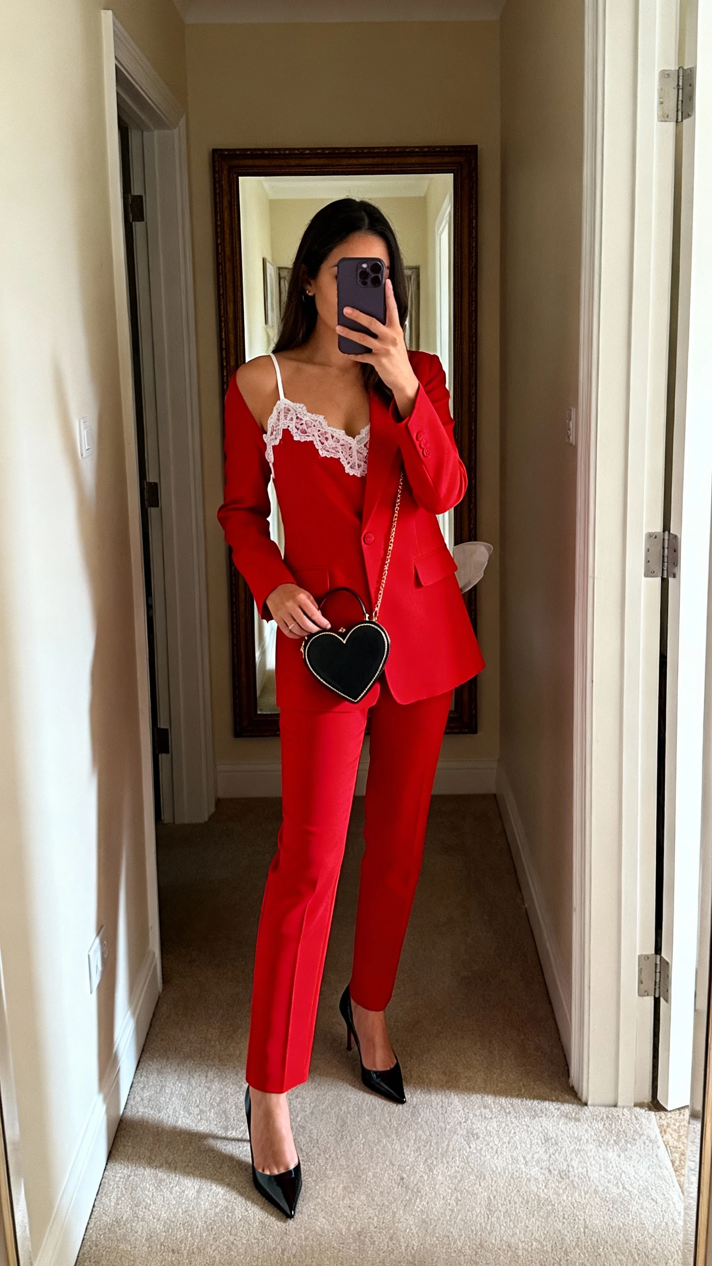 iPhone mirror selfie of a woman wearing a red power suit with a sweet detail (lace-trim camisole), pointed heels, and a heart-shaped mini bag, face not visible, full-length mirror in entryway, natural indoor lighting, iPhone photo quality.