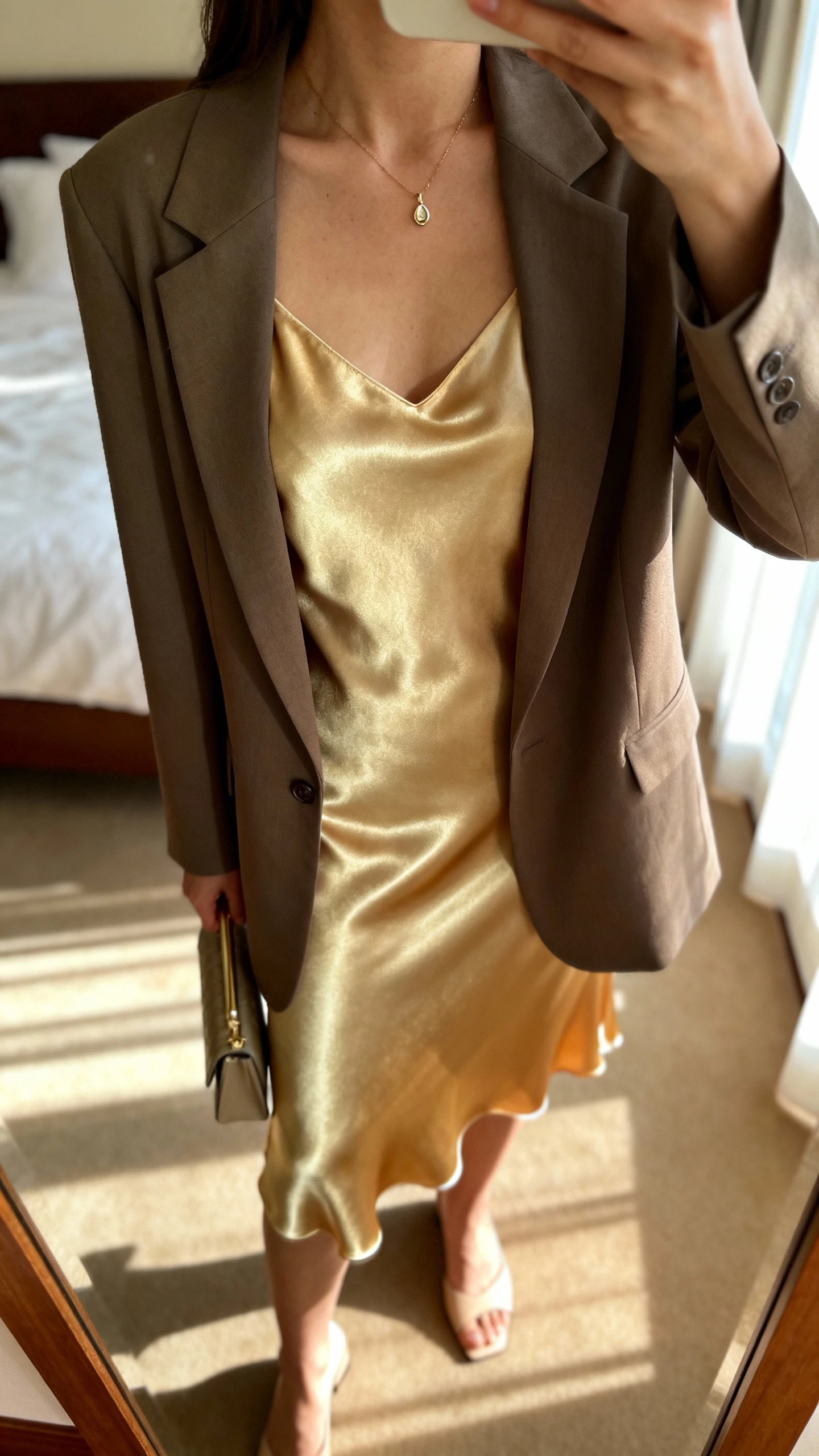 iPhone mirror selfie of a woman wearing a silk slip dress with a tailored blazer, delicate pendant necklace, minimalist heels, and a small clutch, face not visible, cropped at neck, bedroom mirror, soft morning window light, iPhone photo quality.
