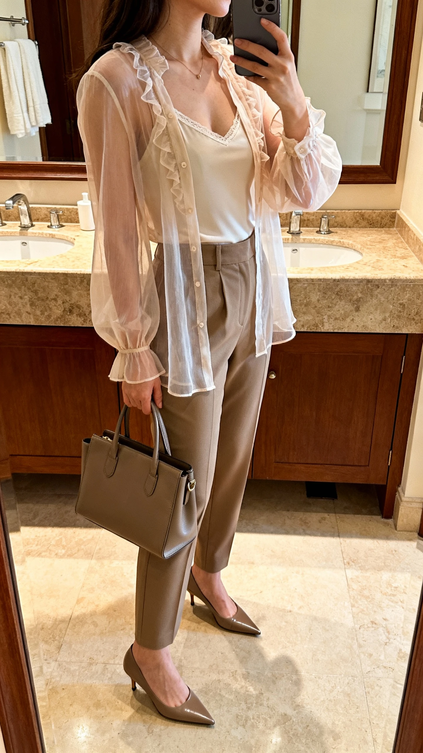 iPhone mirror selfie side angle of a woman in a romantic sheer blouse with a camisole underneath, high-waist trousers, pointed-toe pumps, and a structured tote, face not visible, bathroom mirror, indoor warm lighting, iPhone photo quality.