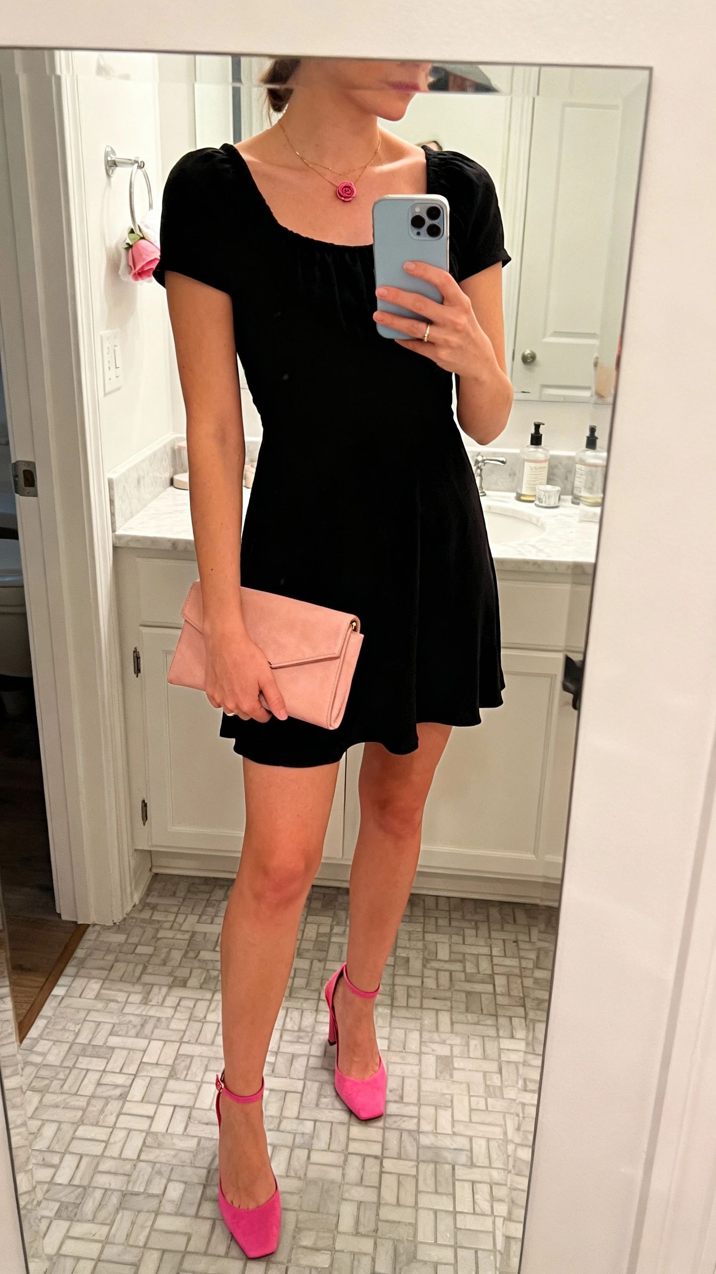 iPhone side-angle mirror selfie of a woman in a little black dress with rosy accents (rose-pink heels and a blush-toned clutch), subtle rose jewelry, face not visible, cropped at neck, bathroom mirror, soft overhead light, iPhone photo quality.