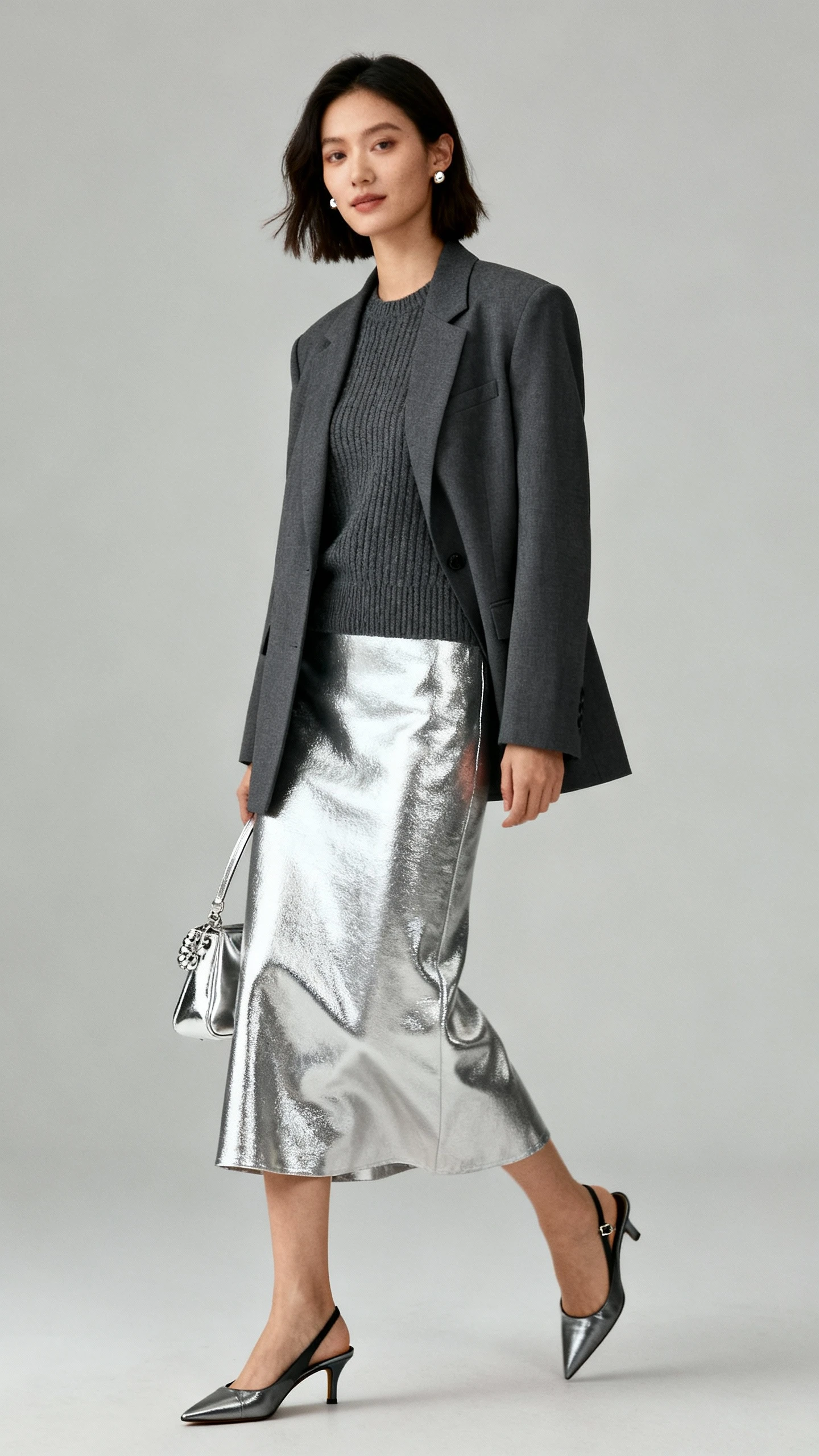 Editorial studio image of a woman in a silver metallic midi skirt with a fine-gauge charcoal knit, a tailored blazer, slingback heels, a metallic-accent bag, and simple studs; neutral gray background, soft studio lighting, subtle movement, clean composition.