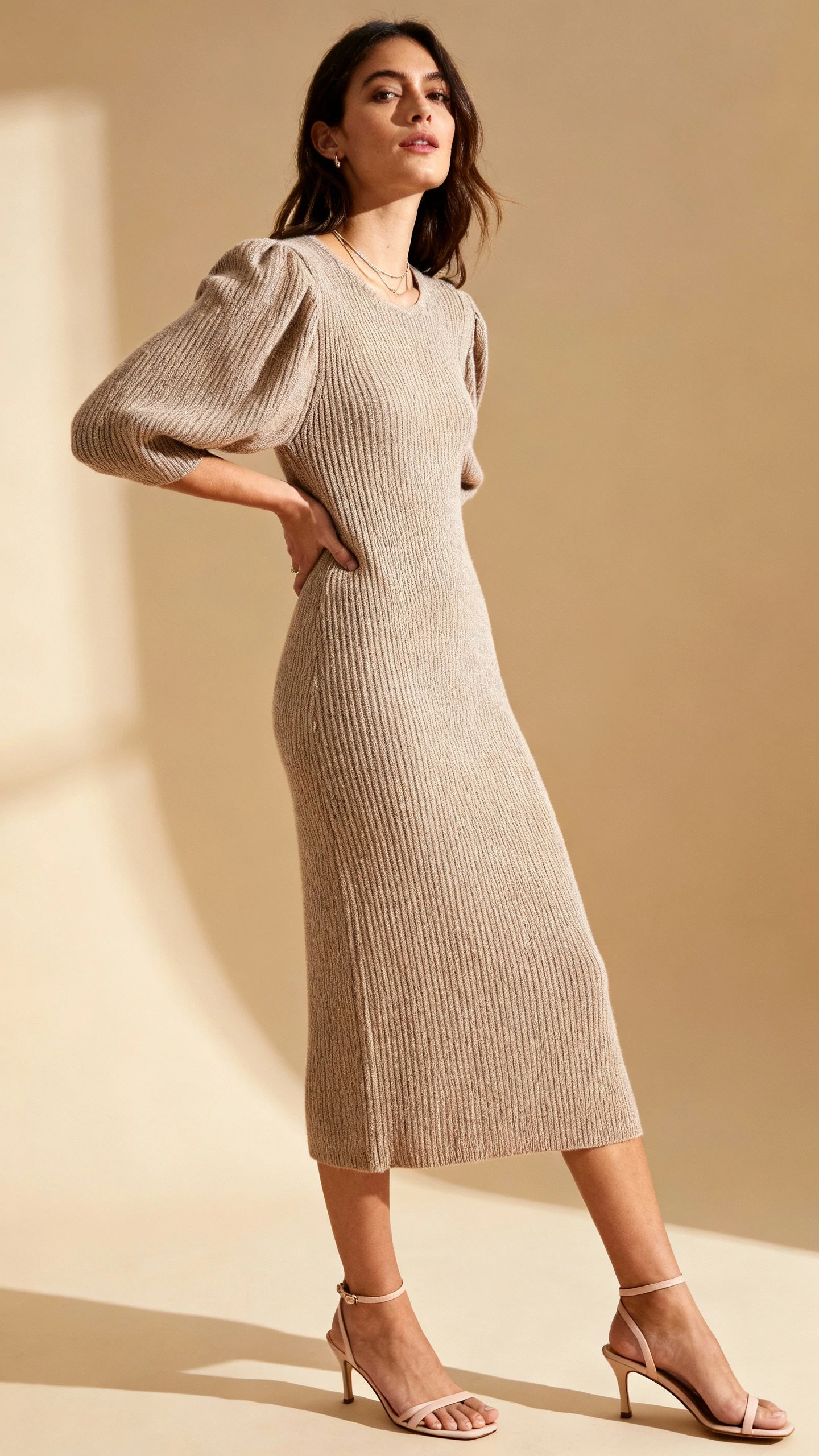 Fashion portrait of a woman wearing an elevated knit midi dress with gentle volume or subtle ribbing, paired with delicate heeled sandals and minimal jewelry, tan background, studio lighting, quiet drama pose, clean composition, editorial feel.