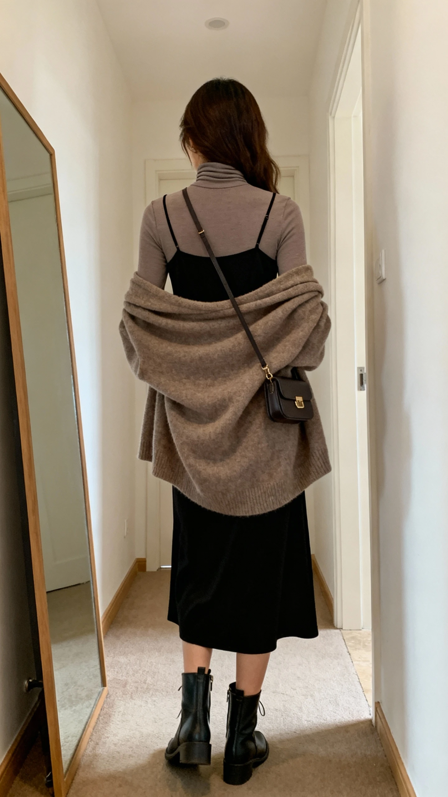 iPhone back view of a woman layering a black slip dress over a thin turtleneck with an oversized cardigan draped over shoulders, ankle boots, small crossbody, face not visible, hallway mirror, gentle indoor lighting, iPhone photo quality.