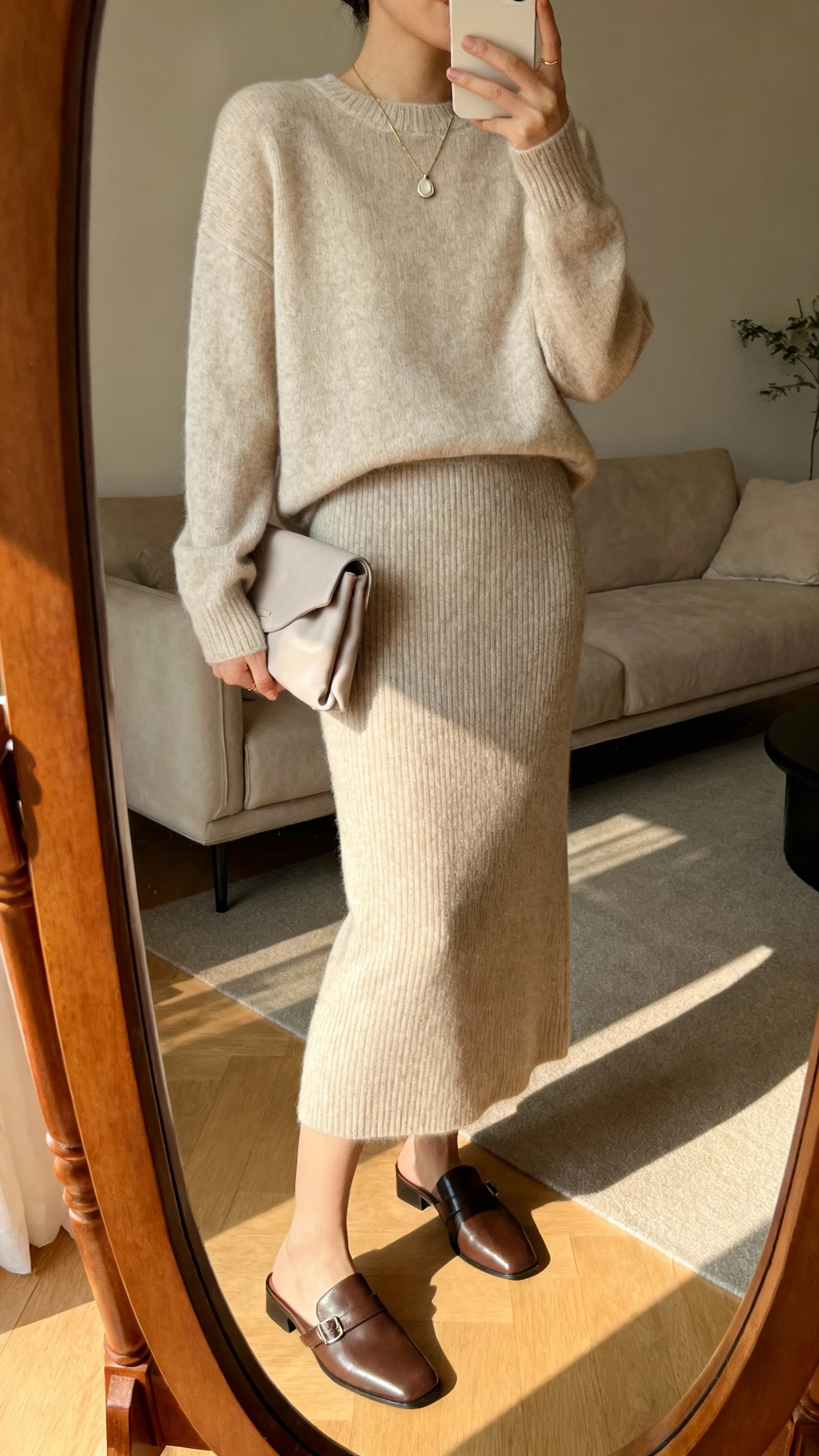 iPhone mirror selfie of a woman in a luxe matching knit set (fine-gauge sweater and midi knit skirt) with sleek leather mules, simple pendant necklace, soft clutch, face not visible, cropped at neck, living room mirror, warm afternoon light, iPhone photo quality.