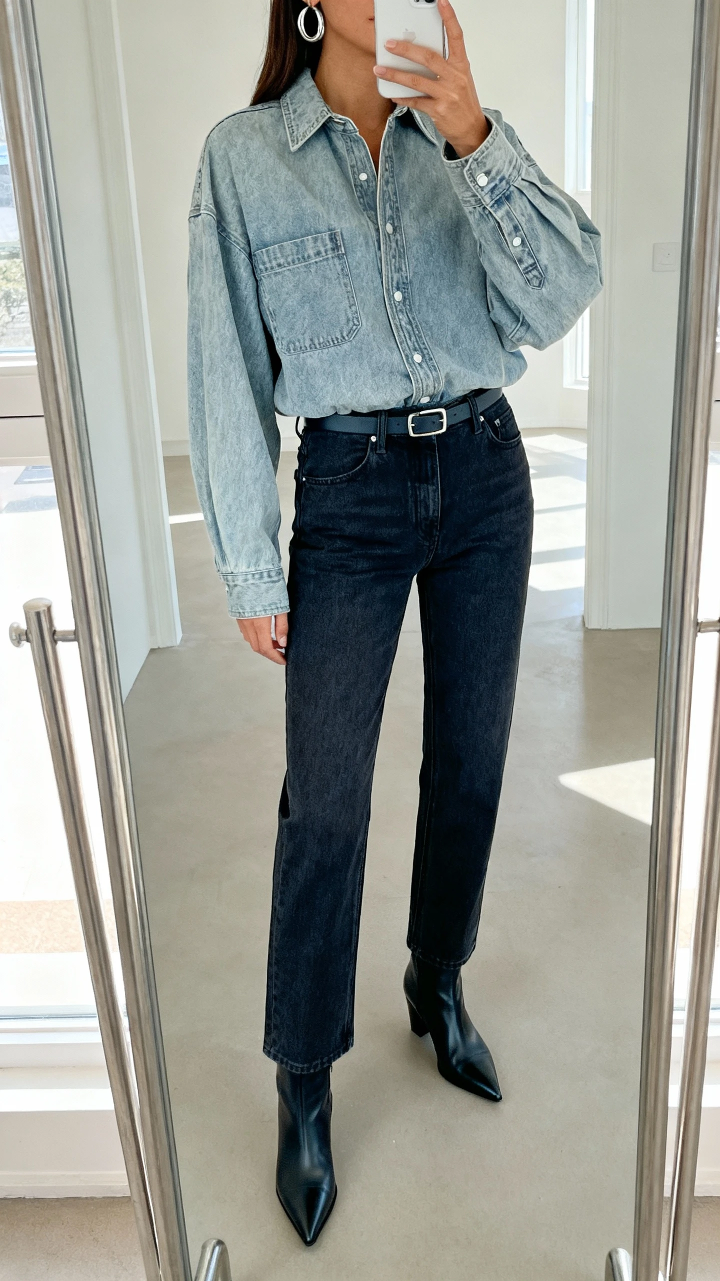 iPhone mirror selfie of a woman in modern denim-on-denim: light-wash oversized denim shirt tucked into darker high-rise straight jeans, pointed ankle boots, slim belt, silver hoops, face not visible, cropped at neck, full-length mirror, natural daylight, iPhone photo quality.