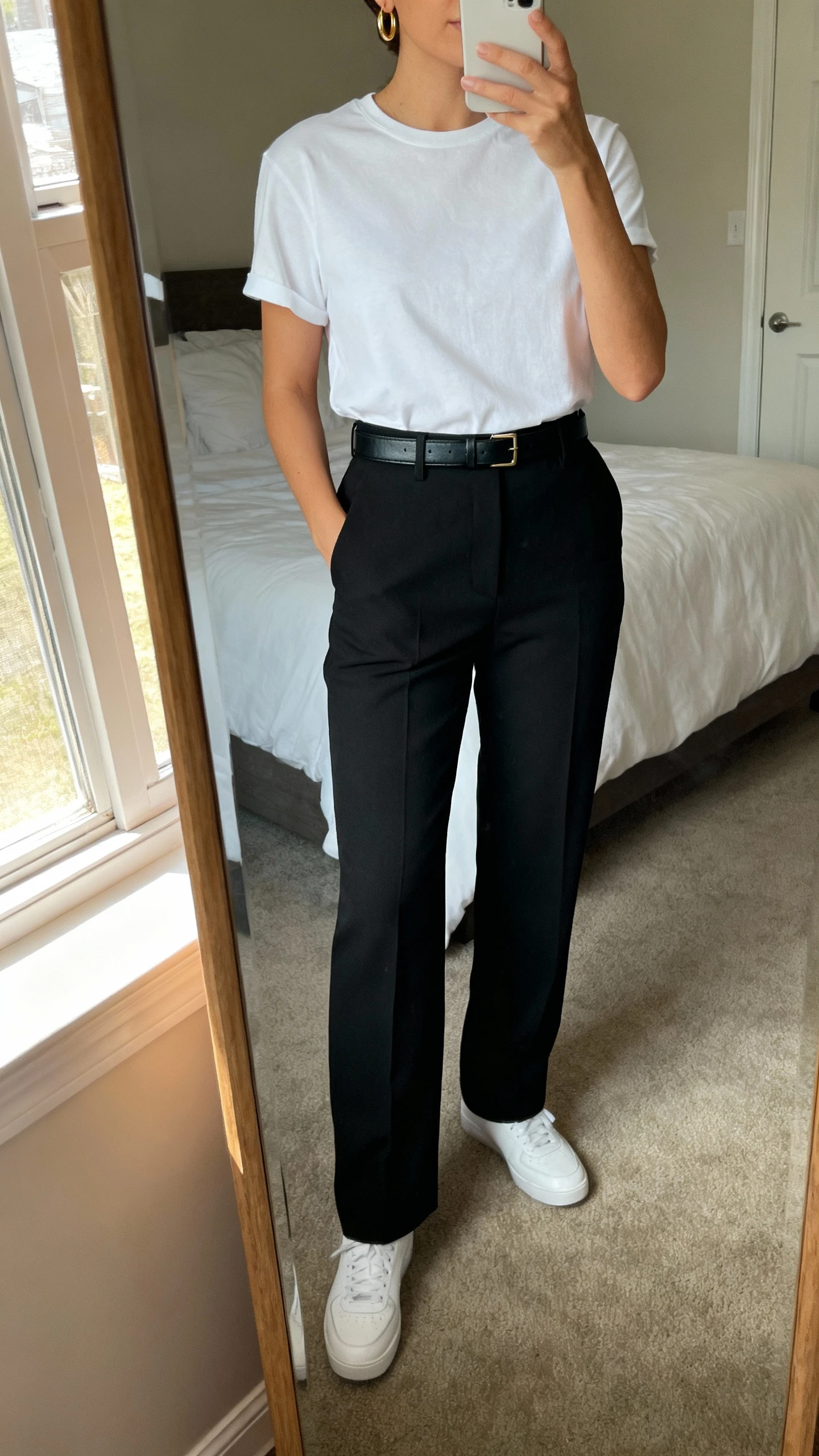 iPhone mirror selfie of a woman wearing an elevated white tee tucked into high-waisted tailored black trousers with a slim leather belt, minimalist white sneakers, delicate gold hoops, face not visible, cropped at neck, bedroom mirror, natural window light, iPhone photo quality.