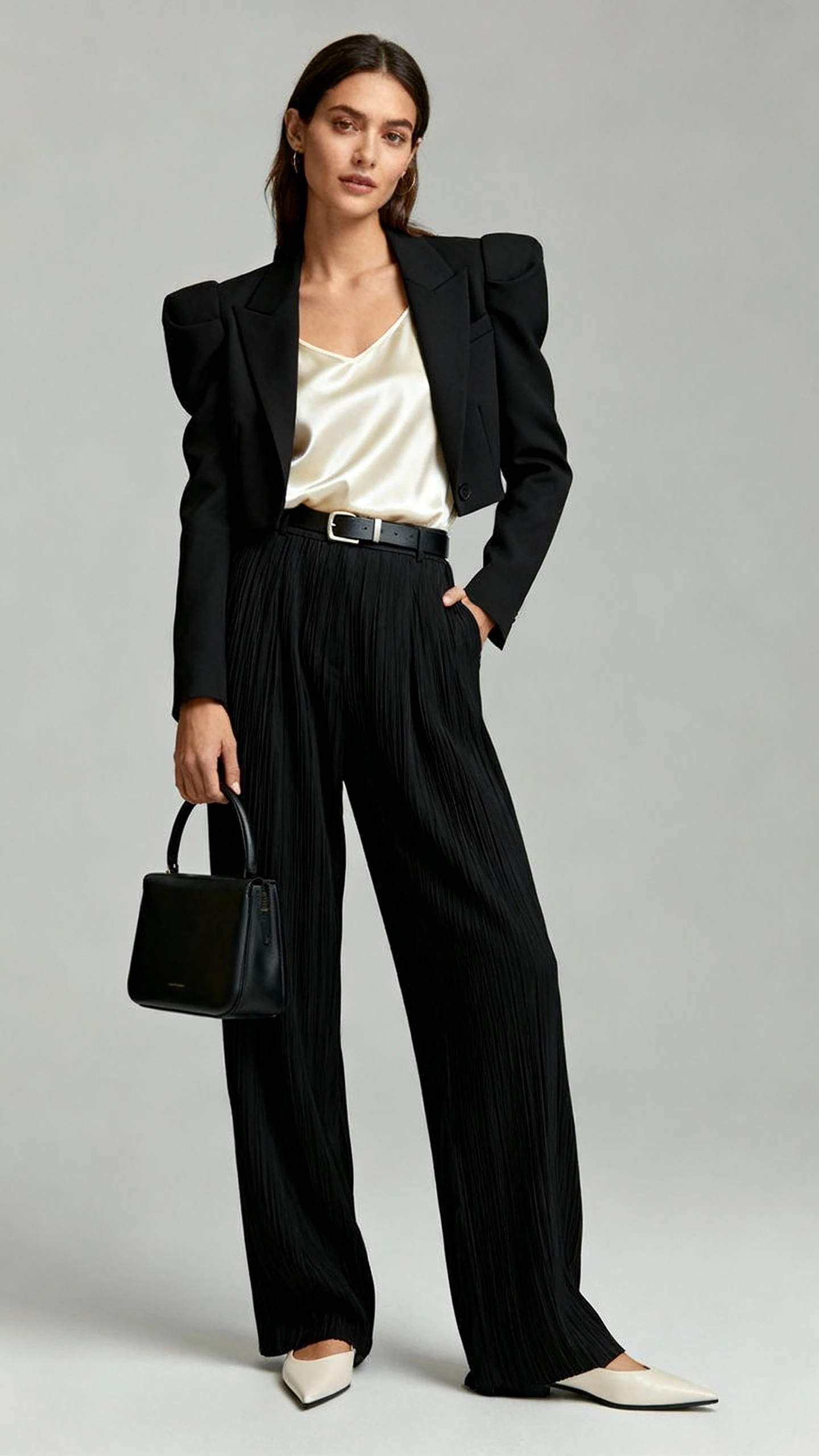 Studio fashion photo of a woman wearing a black sculpted blazer with padded shoulders, high-waisted relaxed pleated trousers, a silky cream tank, pointed-toe loafers, a minimal belt, and a structured top-handle bag; neutral gray background, studio lighting, professional natural pose, clean composition, high quality.