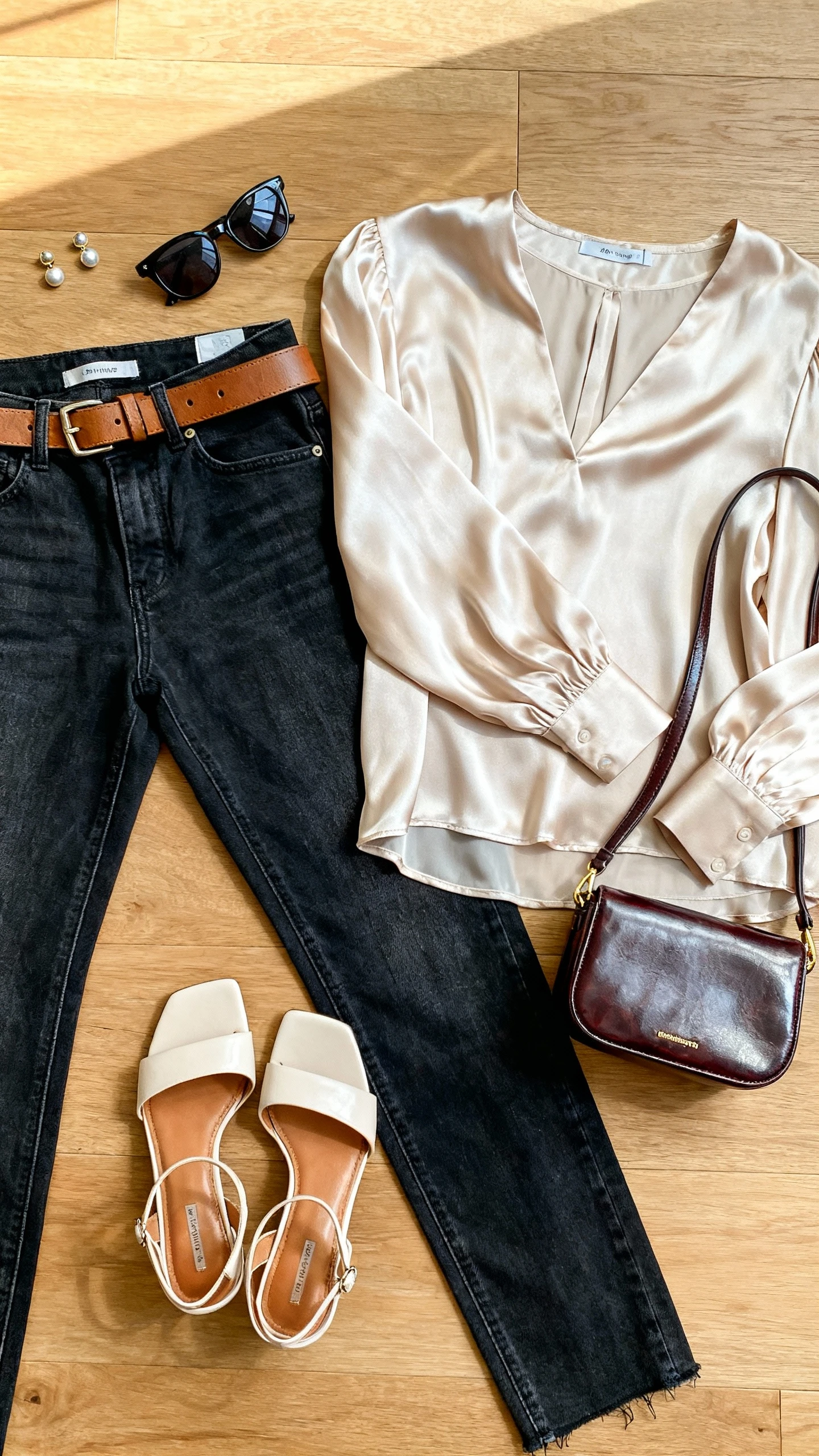 Top-down flat lay of dark-wash jeans with an elevated top (silk blouse), heeled sandals, belt, and a polished mini bag on a wooden floor; include stud earrings and sunglasses, bright natural light, no person visible, organized composition, iPhone photo aesthetic.