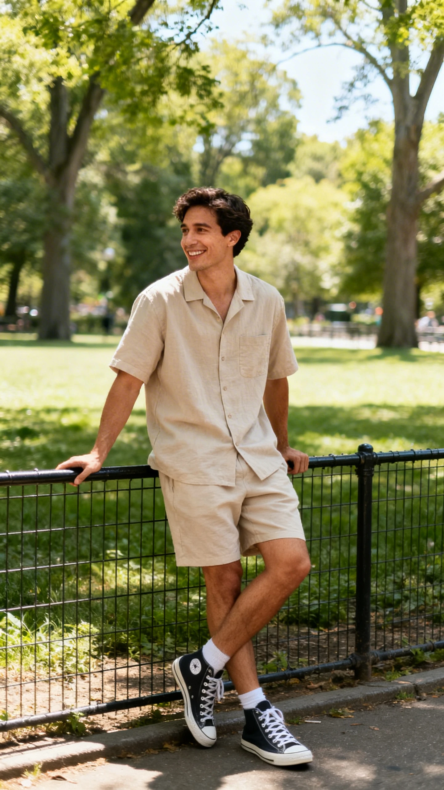 A man wearing a matching lightweight co-ord set (short-sleeve shirt and shorts) with canvas sneakers, leaning on a park fence and smiling mid-conversation, casual iPhone photo style, sunny day, outdoor setting.
