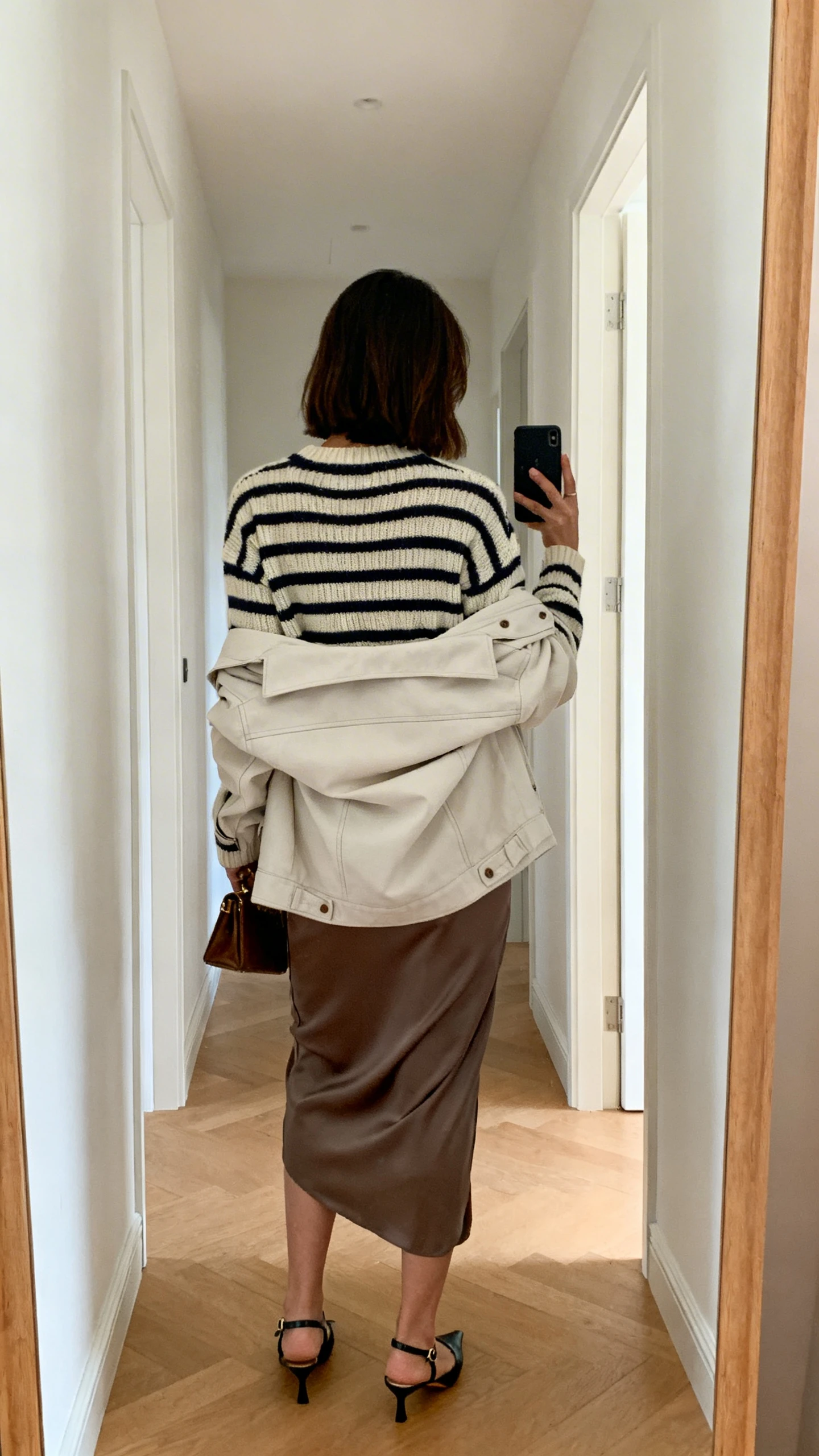 iPhone back-view hallway mirror selfie of a woman wearing a striped knit sweater, bias-cut slip skirt, and a light jacket draped over shoulders, with slingback flats and a small clutch, face not visible, natural daylight, iPhone photo quality