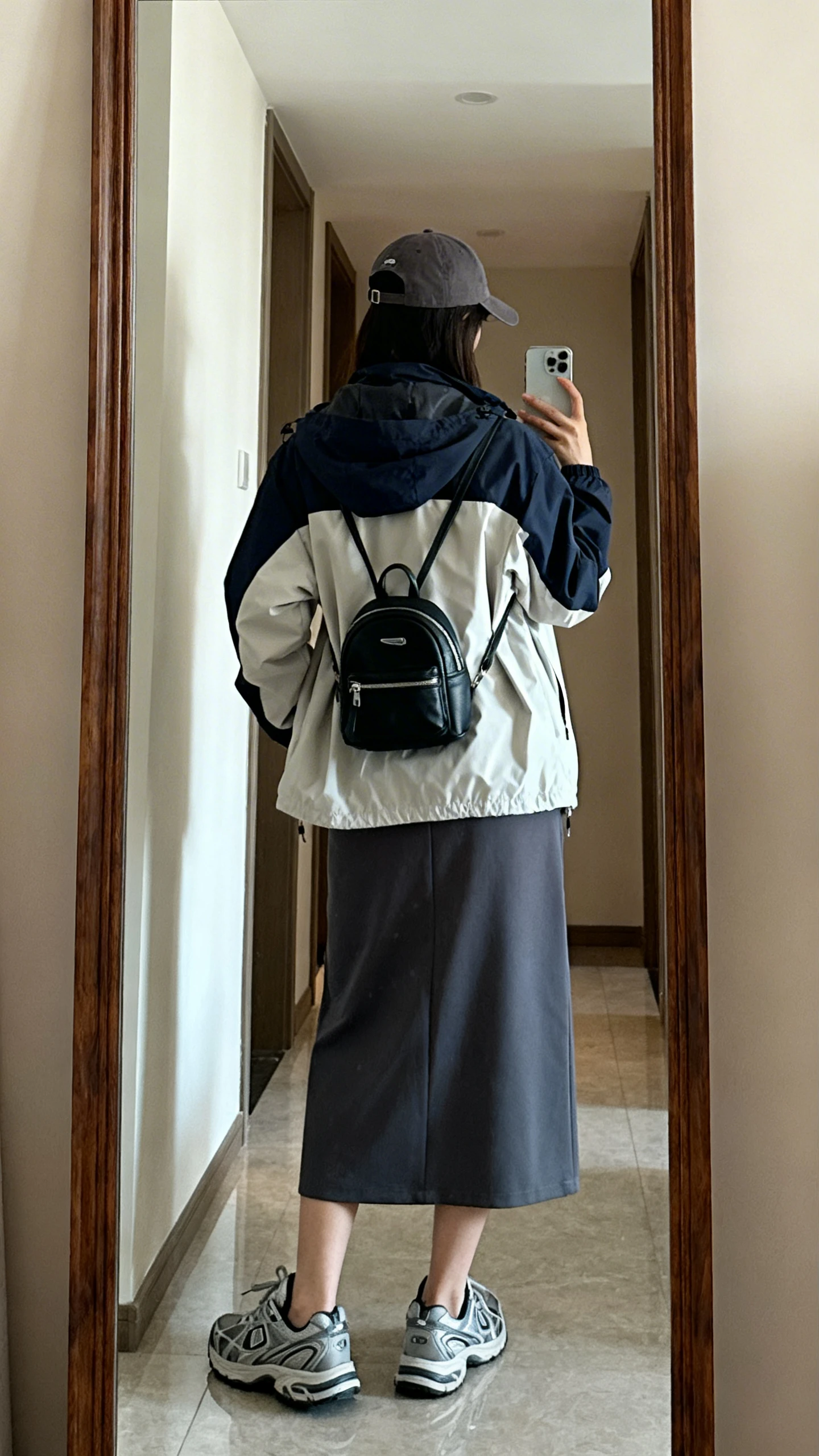 iPhone back-view mirror selfie of a woman wearing a sporty windbreaker layered over a midi skirt, sleek trainers, baseball cap, and mini backpack, face not visible, hallway full-length mirror, natural daylight, iPhone photo quality