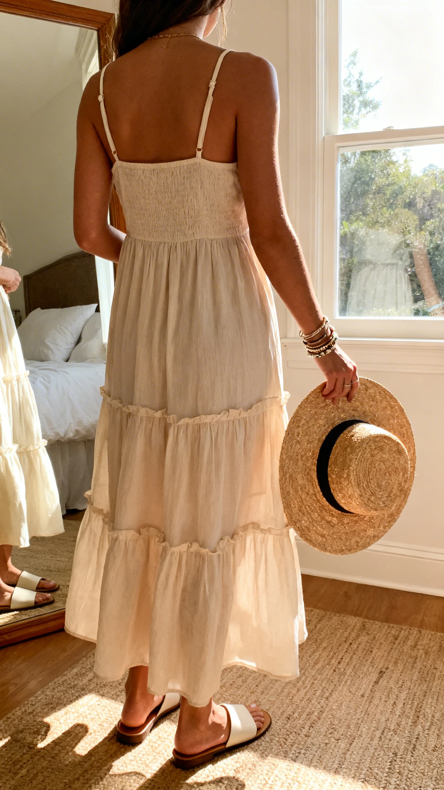 iPhone back view of a woman wearing a breezy tiered sundress with thin straps, flat slide sandals, a straw sun hat in hand, and layered bracelets, face not visible, standing by a bedroom mirror, warm afternoon window light, iPhone photo quality.