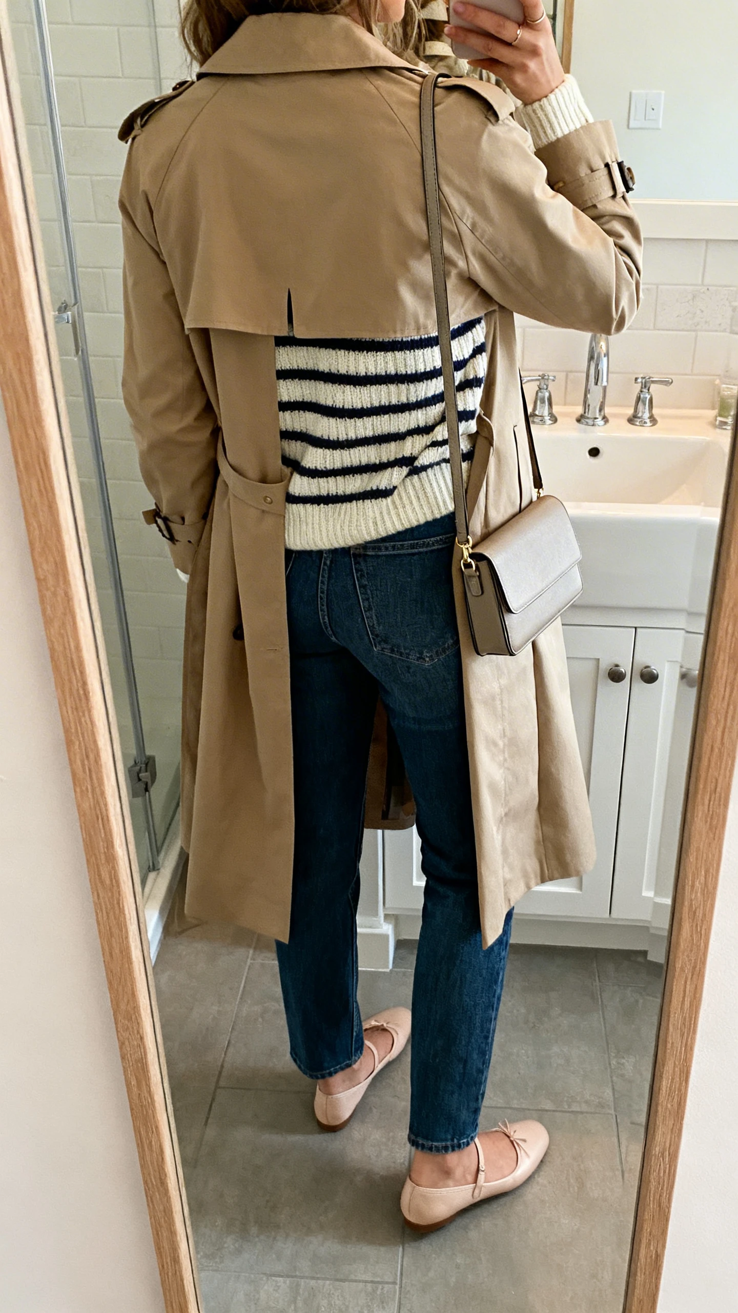 iPhone back-view selfie of a woman wearing a classic trench coat over a striped knit sweater, straight jeans, ballet flats, and a slim crossbody; face not visible, bathroom mirror, soft indoor lighting, iPhone photo quality.