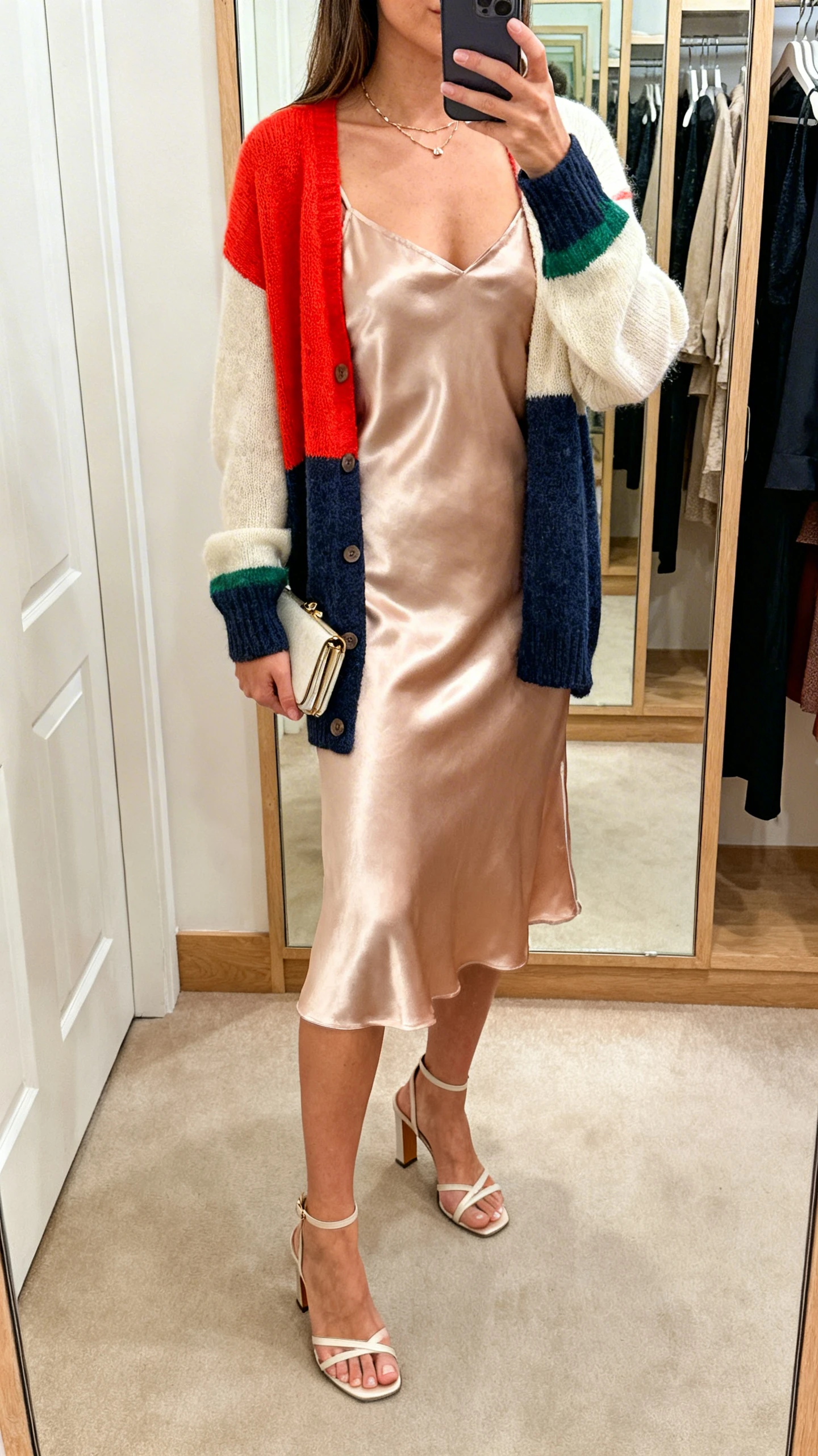 iPhone mirror selfie of a woman in a color-pop cardigan layered over a satin slip dress, with delicate necklace, heeled sandals, and a small clutch; face not visible, cropped at shoulders, dressing-area mirror with warm indoor light, iPhone photo quality.