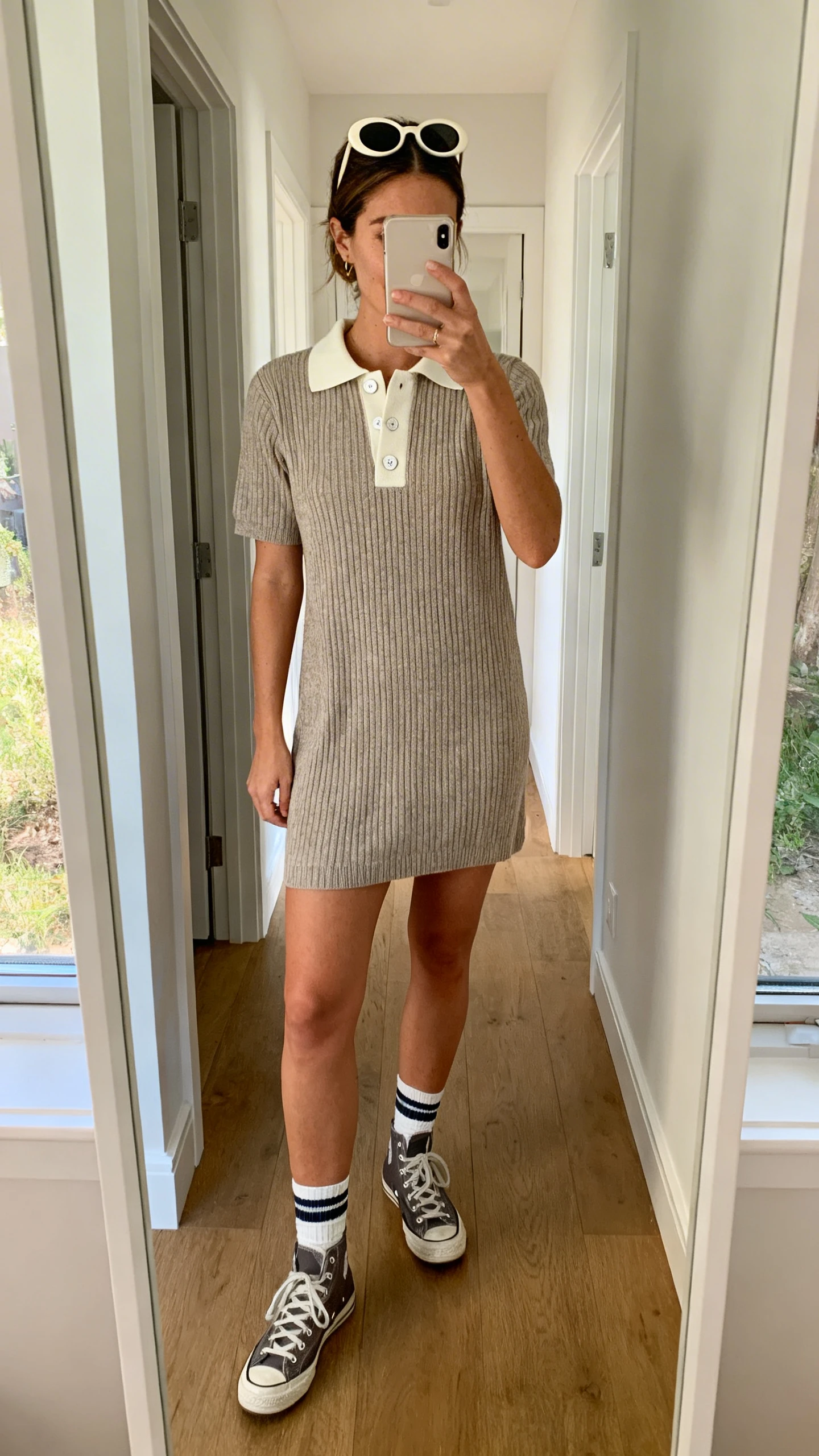 iPhone mirror selfie of a woman in a knit polo dress with a collar and buttons, retro sneakers, crew socks, and oval sunglasses pushed into hair, face not visible, cropped at shoulders, full-length hallway mirror, natural daylight, iPhone photo quality.