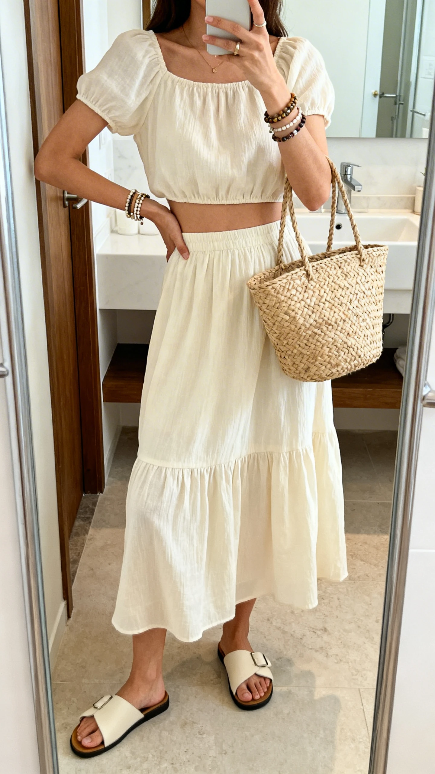 iPhone mirror selfie of a woman in a matching co-ord set: cropped top and breezy midi skirt, with slide sandals, layered bracelets, and a basket tote, face not visible, bathroom mirror, bright indoor lighting, iPhone photo quality.