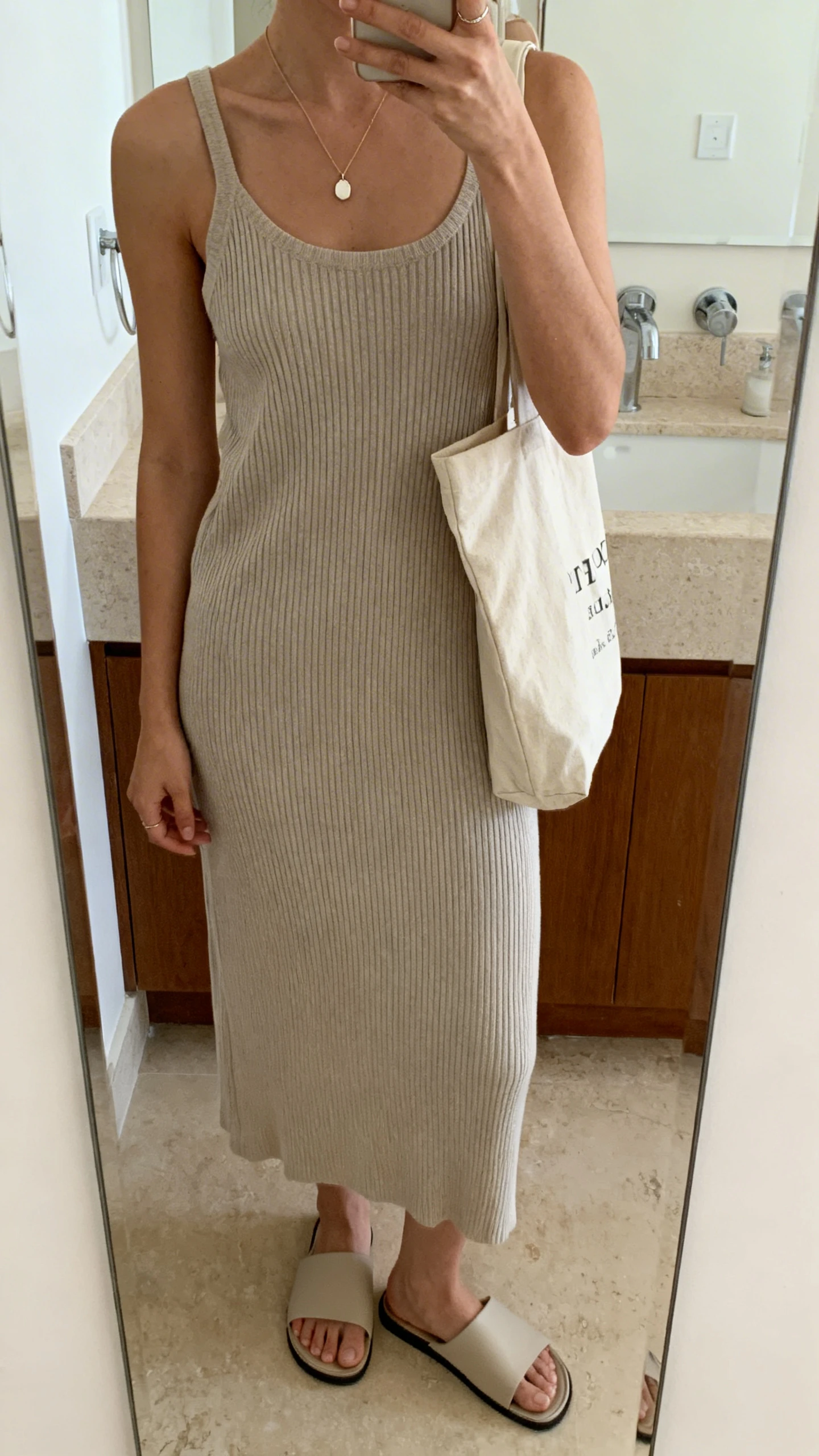 iPhone mirror selfie of a woman in a ribbed knit dress styled with minimalist slide sandals, delicate pendant necklace, and a canvas tote, face not visible, cropped at shoulders, bathroom mirror, soft indoor lighting, iPhone photo quality.