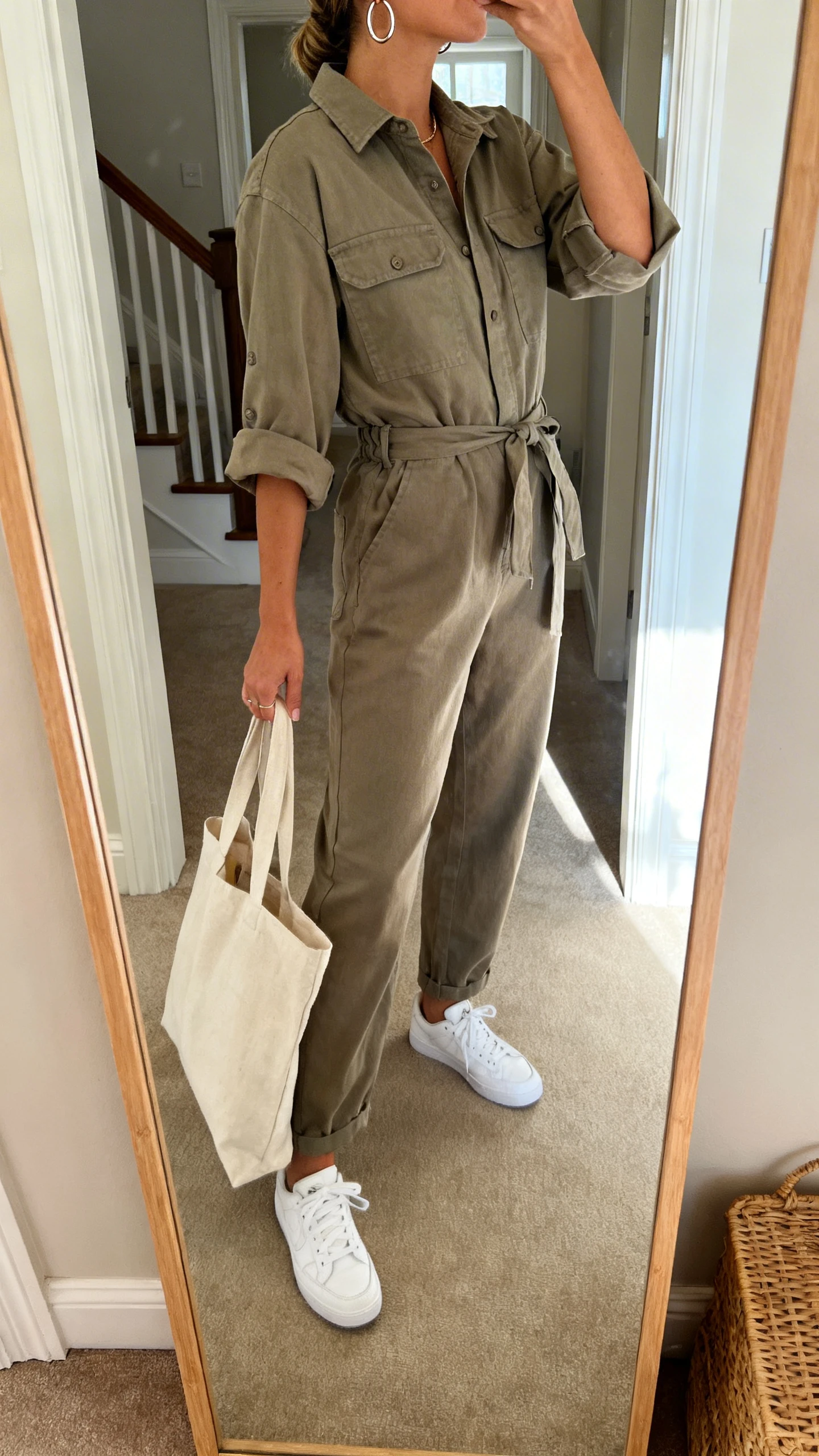 iPhone mirror selfie of a woman in a utility jumpsuit with rolled sleeves, cinched waist, white sneakers, hoop earrings, and a canvas tote; face not visible, side angle, entryway full-length mirror, natural daylight, iPhone photo quality.