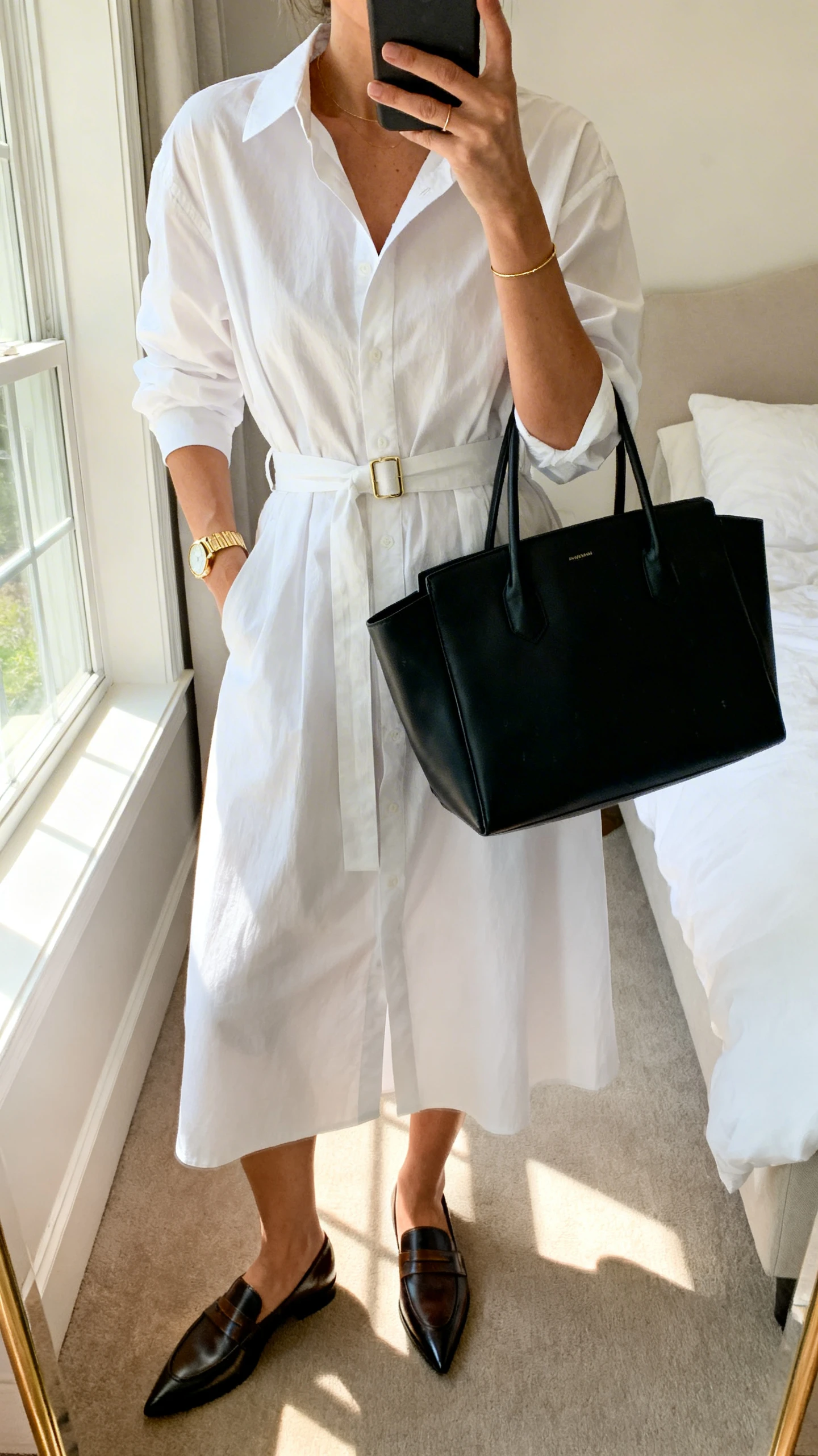 iPhone mirror selfie of a woman wearing a crisp white poplin shirt dress with a belted waist, pointed toe loafers, a structured black tote, and a thin gold watch, face not visible, cropped at neck, bedroom mirror, morning window light, iPhone photo quality