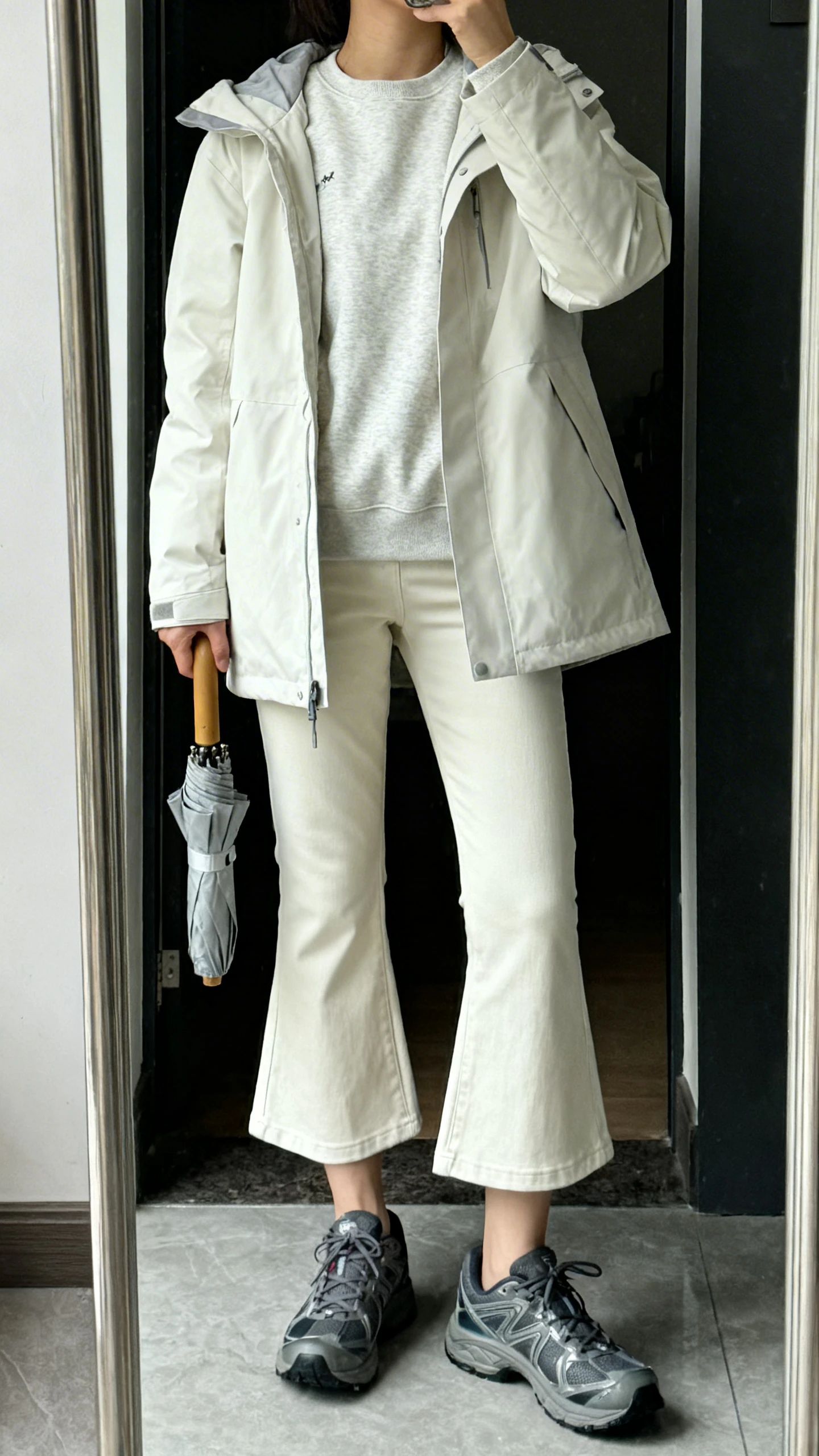 iPhone mirror selfie of a woman wearing a light parka over a crewneck sweatshirt with cropped kick-flare pants, waterproof sneakers, and a compact umbrella in hand, face not visible, cropped at neck, entryway full-length mirror, overcast natural light, iPhone photo quality