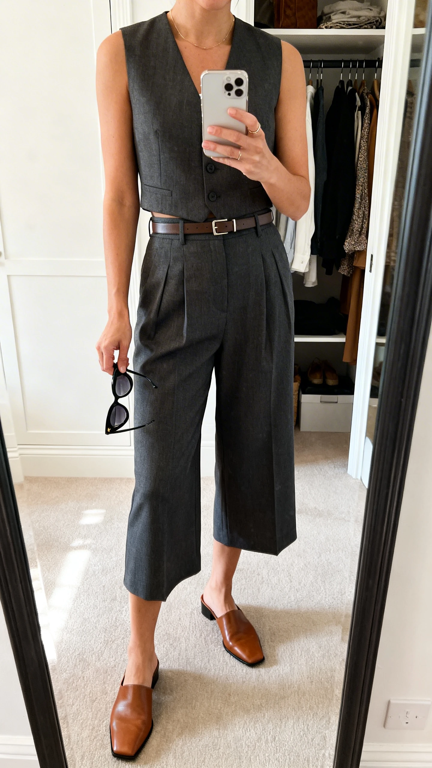 iPhone mirror selfie of a woman wearing a tailored vest with long, knee-grazing shorts, leather mules, slim belt, and cat-eye sunglasses in hand, face not visible, cropped at neck, closet mirror, morning natural light, iPhone photo quality.