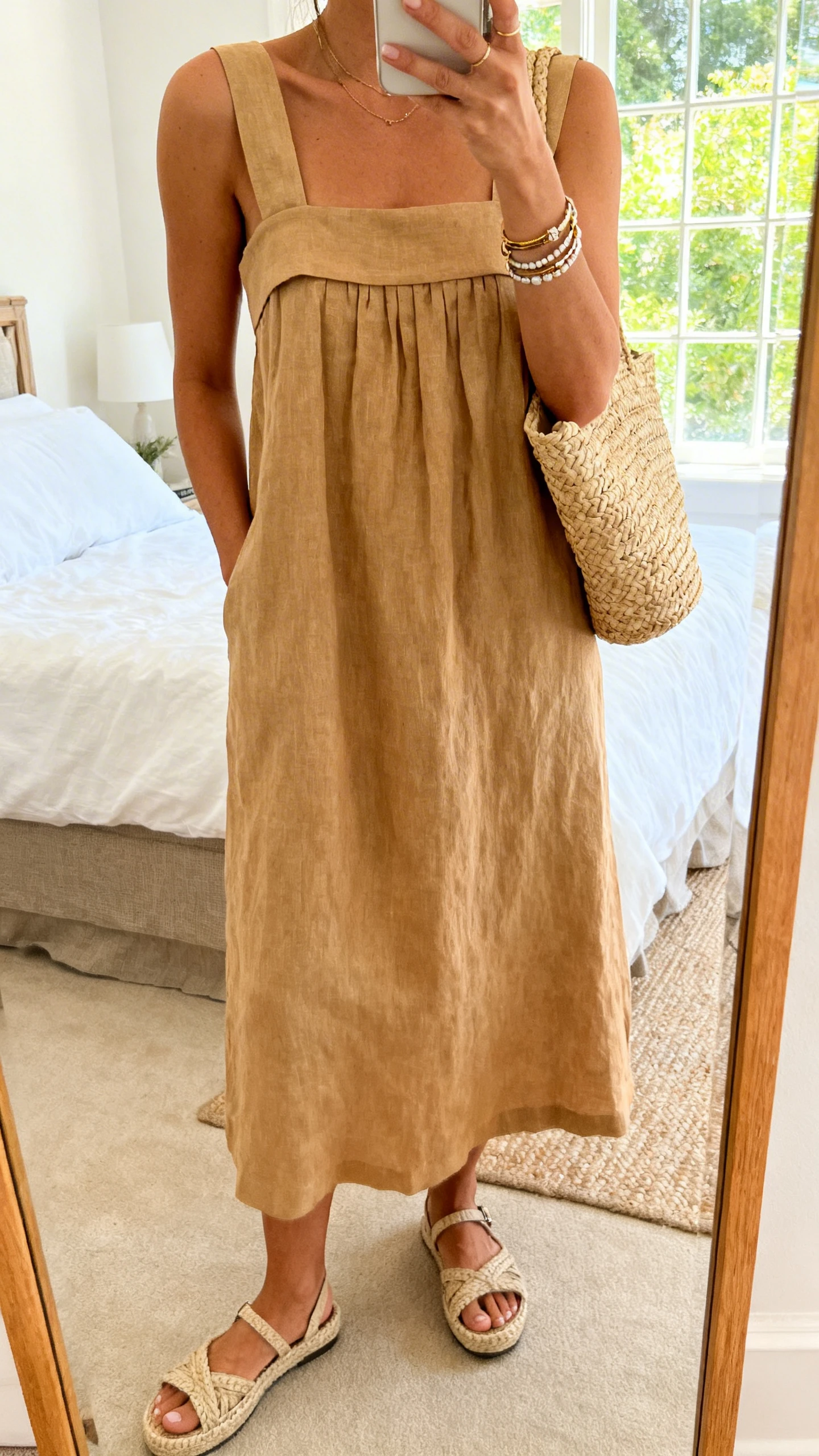 iPhone mirror selfie of a woman wearing a tan linen sundress with wide straps, espadrille sandals, a straw tote, and layered bracelets, face not visible, cropped at shoulders, bedroom mirror, bright afternoon window light, iPhone photo quality