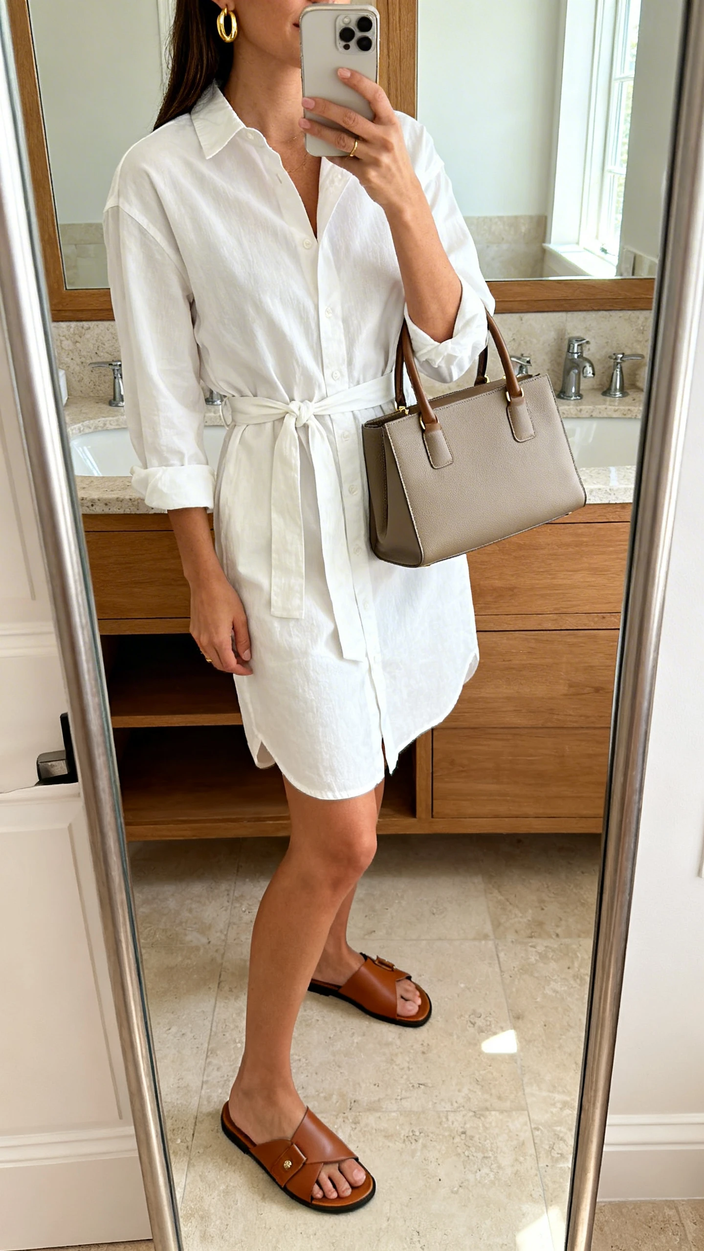 iPhone side-angle mirror selfie of a woman in a crisp cotton shirt dress with a self-tie belt, leather slide sandals, a structured top-handle bag, and gold hoop earrings, face not visible, bathroom mirror, bright indoor lighting, iPhone photo quality.