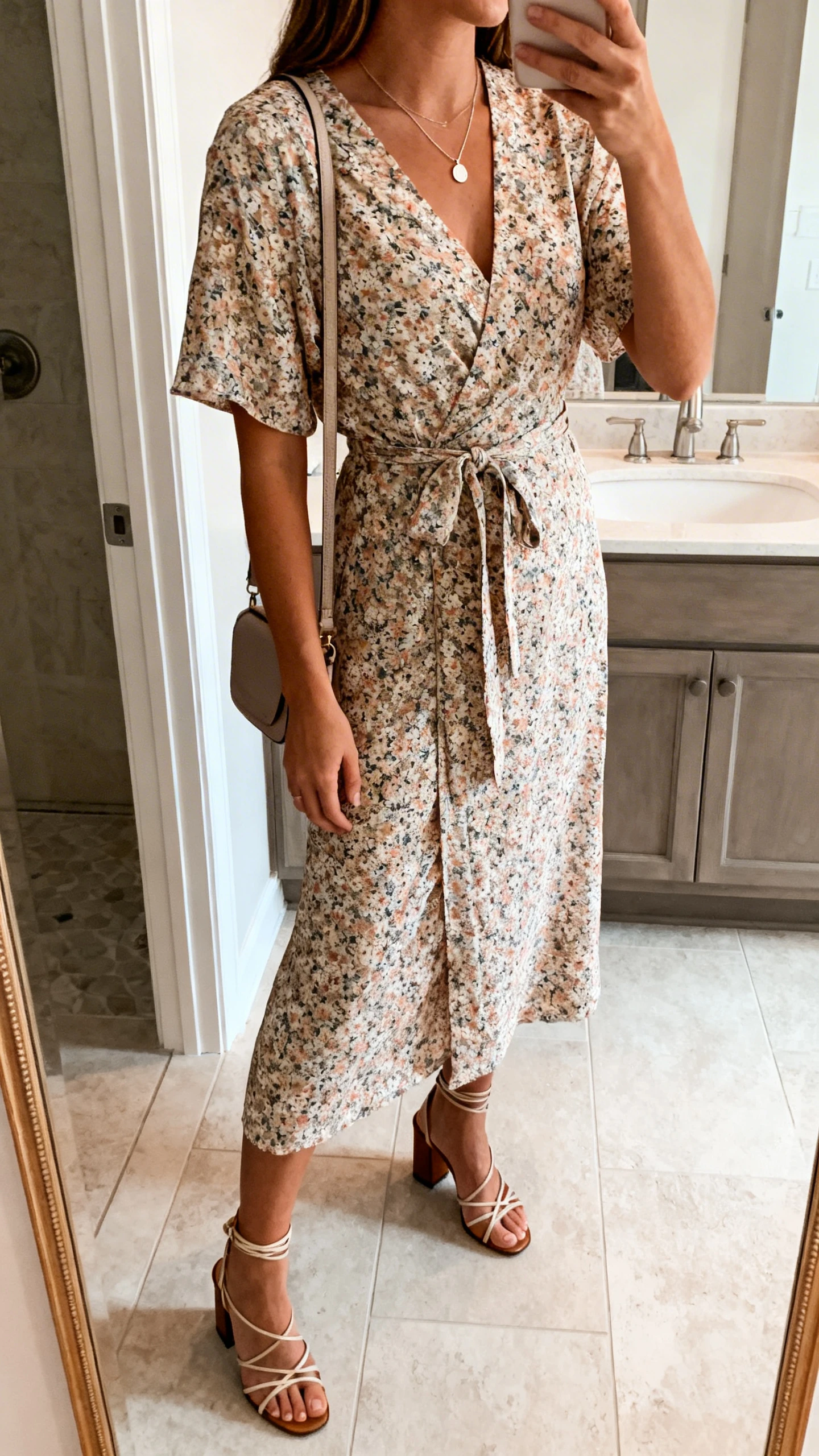 iPhone side-angle mirror selfie of a woman wearing a floral wrap dress with a tie waist, strappy block-heel sandals, a delicate pendant necklace, and a small crossbody bag, face not visible, bathroom mirror, natural indoor lighting, iPhone photo quality.