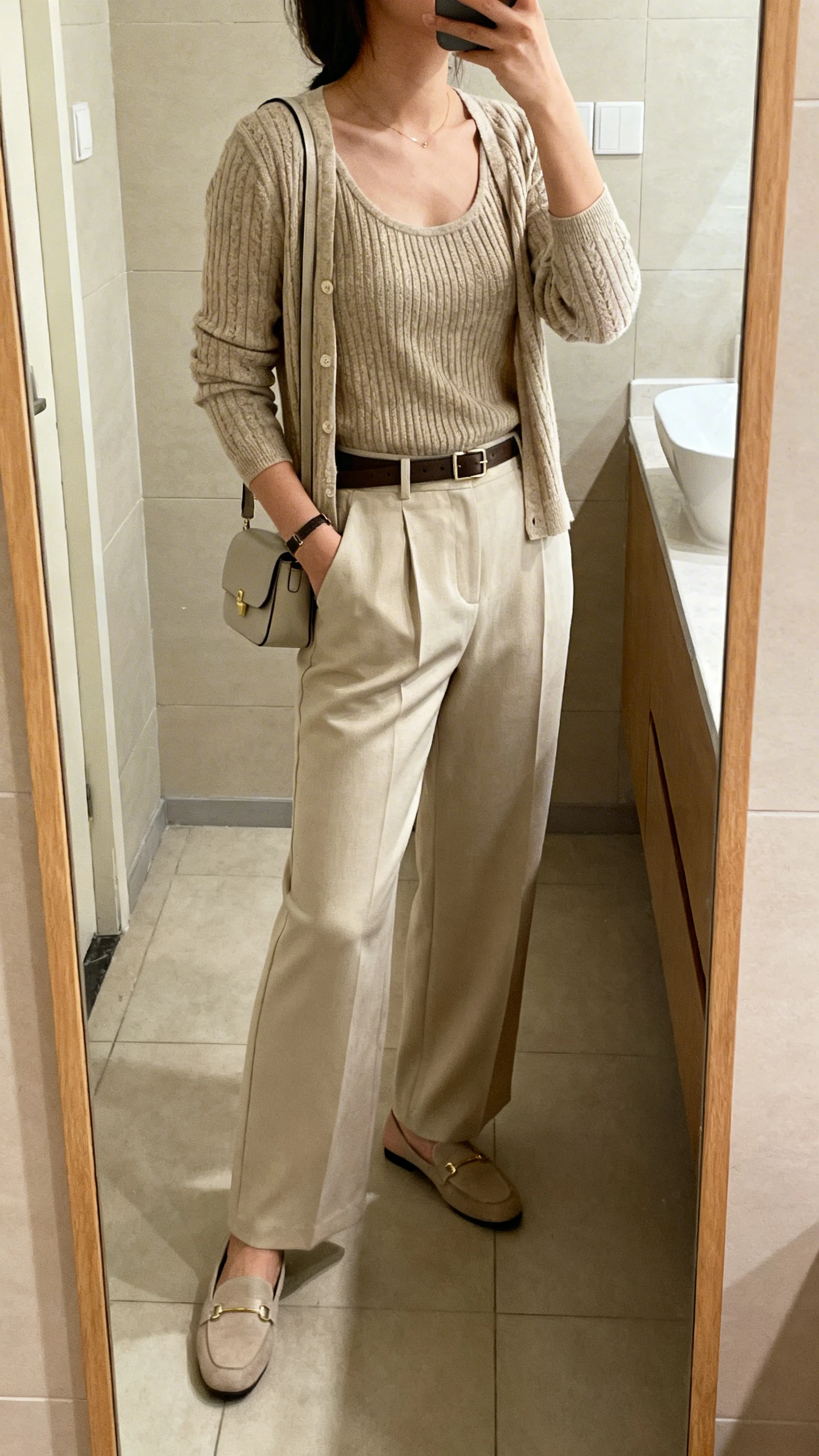 iPhone side-angle mirror selfie of a woman wearing a knit tank with matching cardigan set, high-waisted wide-leg trousers, loafers, slim belt, and small shoulder bag, face not visible, bathroom mirror, soft indoor lighting, iPhone photo quality