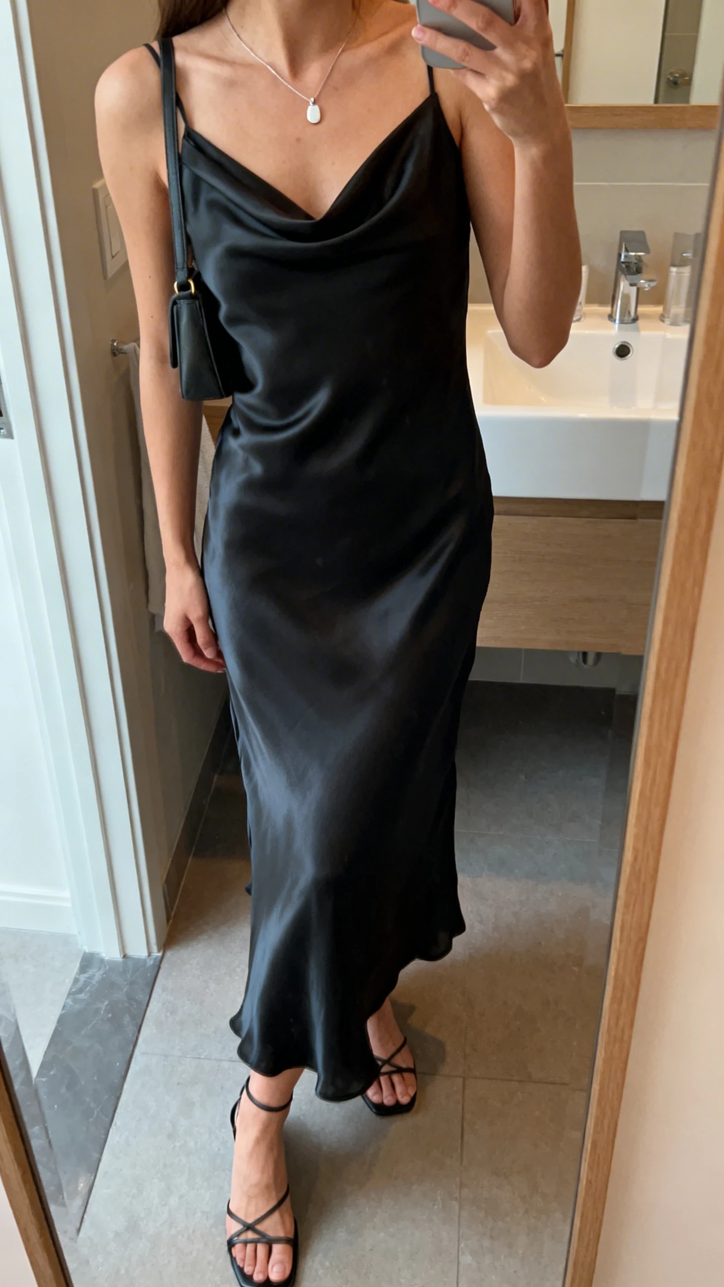 iPhone side-angle mirror selfie of a woman wearing a silky black slip dress with a draped neckline, minimalist strappy heels, a delicate pendant necklace, and a small shoulder bag, face not visible, bathroom mirror, soft indoor lighting, iPhone photo quality