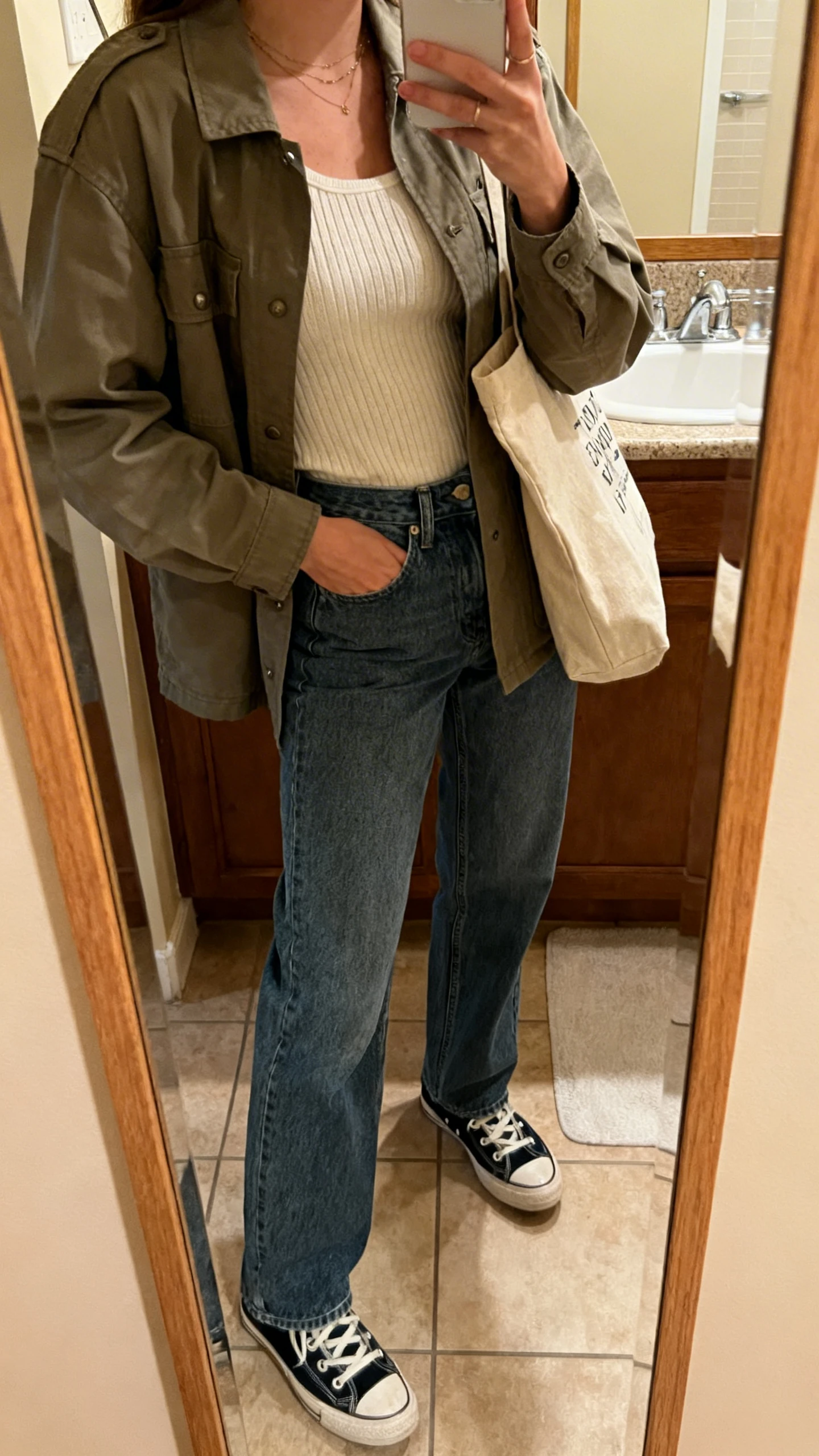 iPhone side-angle mirror selfie of a woman wearing a utility shacket over a soft knit tank with relaxed straight jeans, retro sneakers, and a canvas tote, face not visible, bathroom mirror, warm indoor lighting, iPhone photo quality